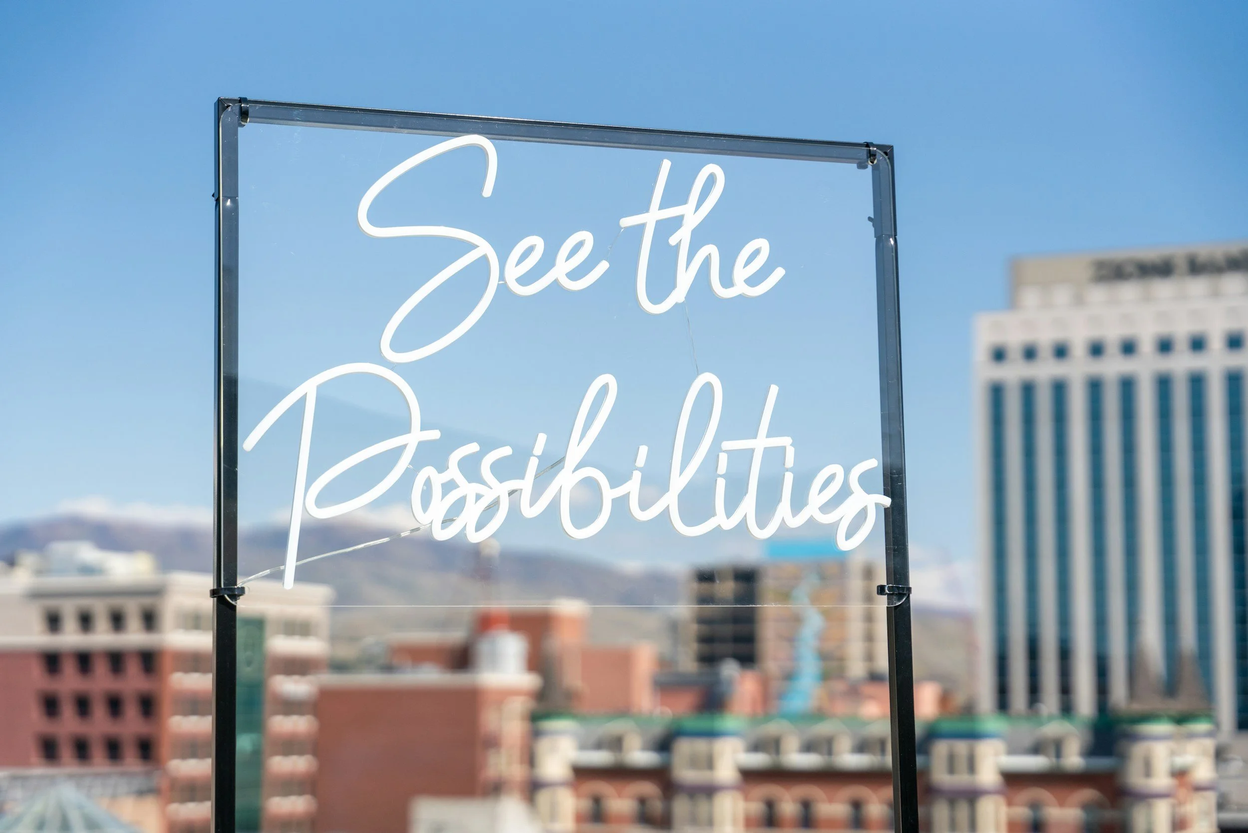 Neon sign on a balcony with a cityscape background, displaying the phrase 'See the Possibilities' in white cursive writing.