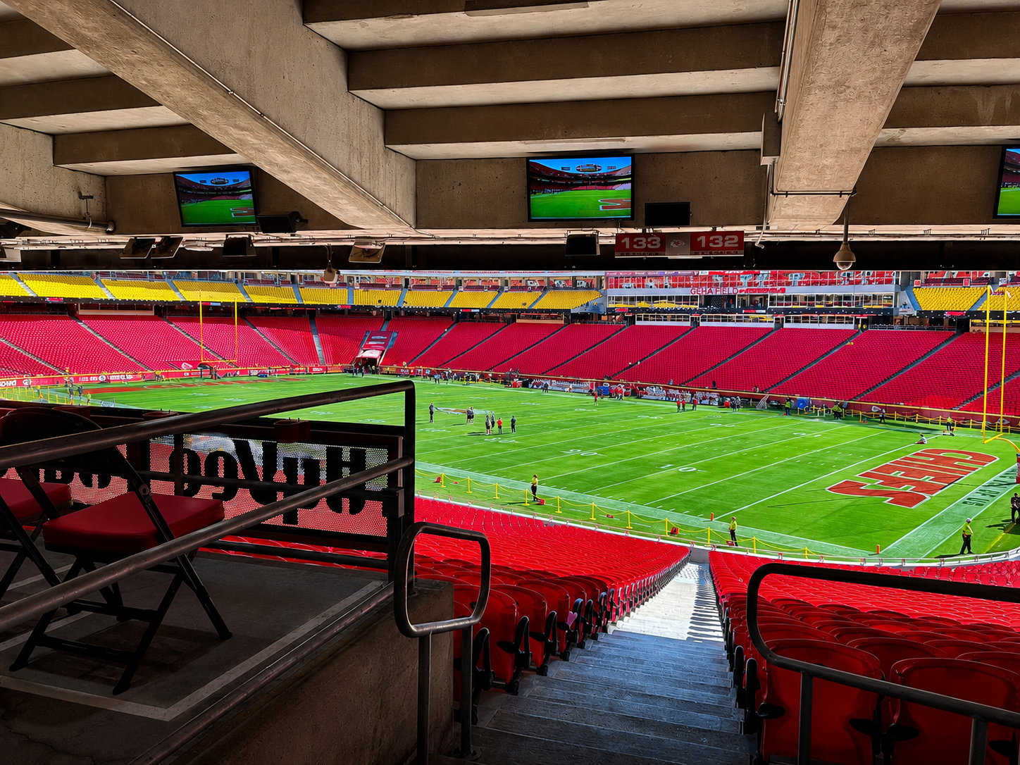 Inside view of an empty football stadium with red and yellow seats, green field, and multiple television screens hanging from the concrete ceiling.