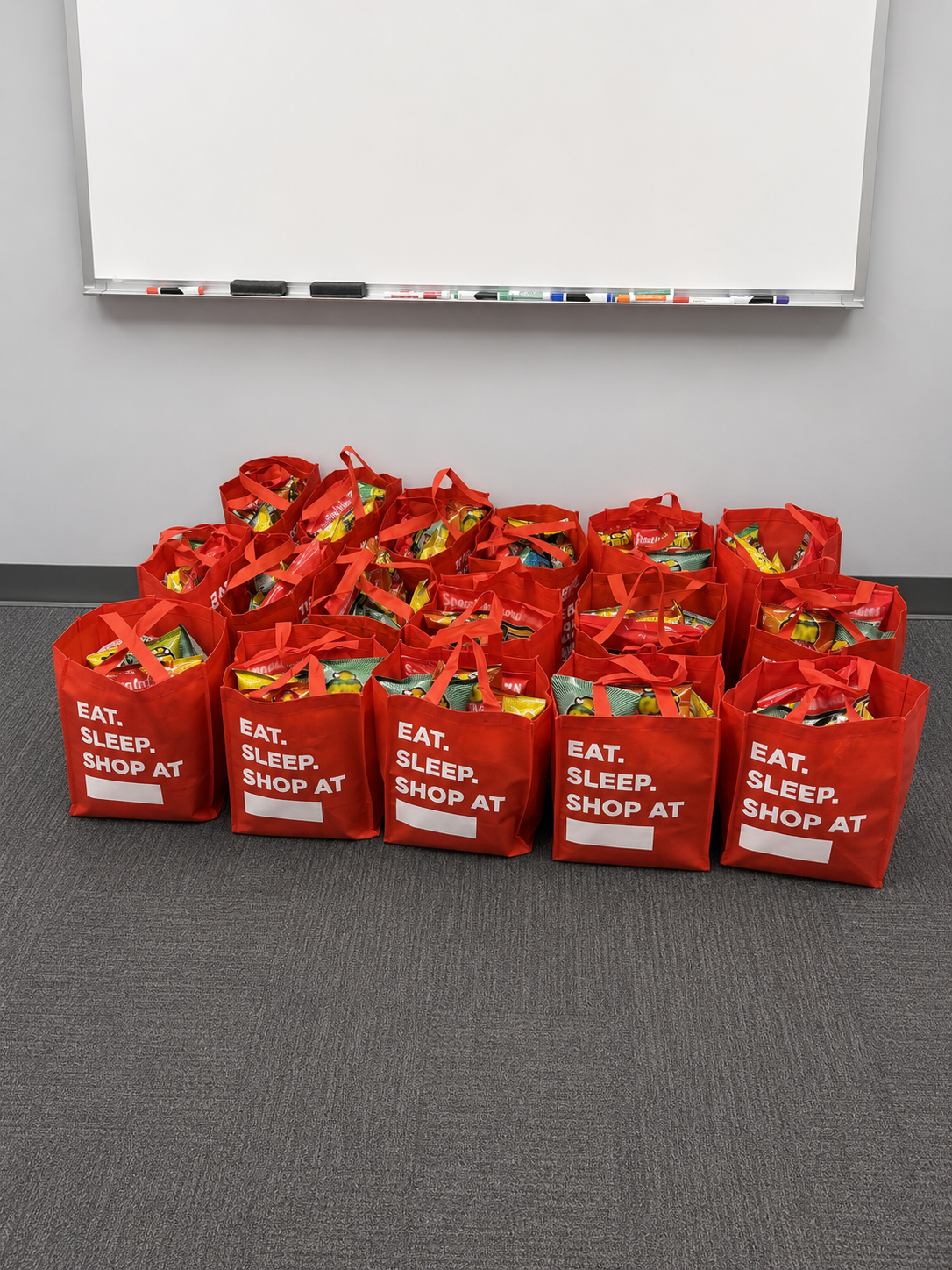 Multiple red shopping bags with snacks placed on the floor in front of a whiteboard in a room.