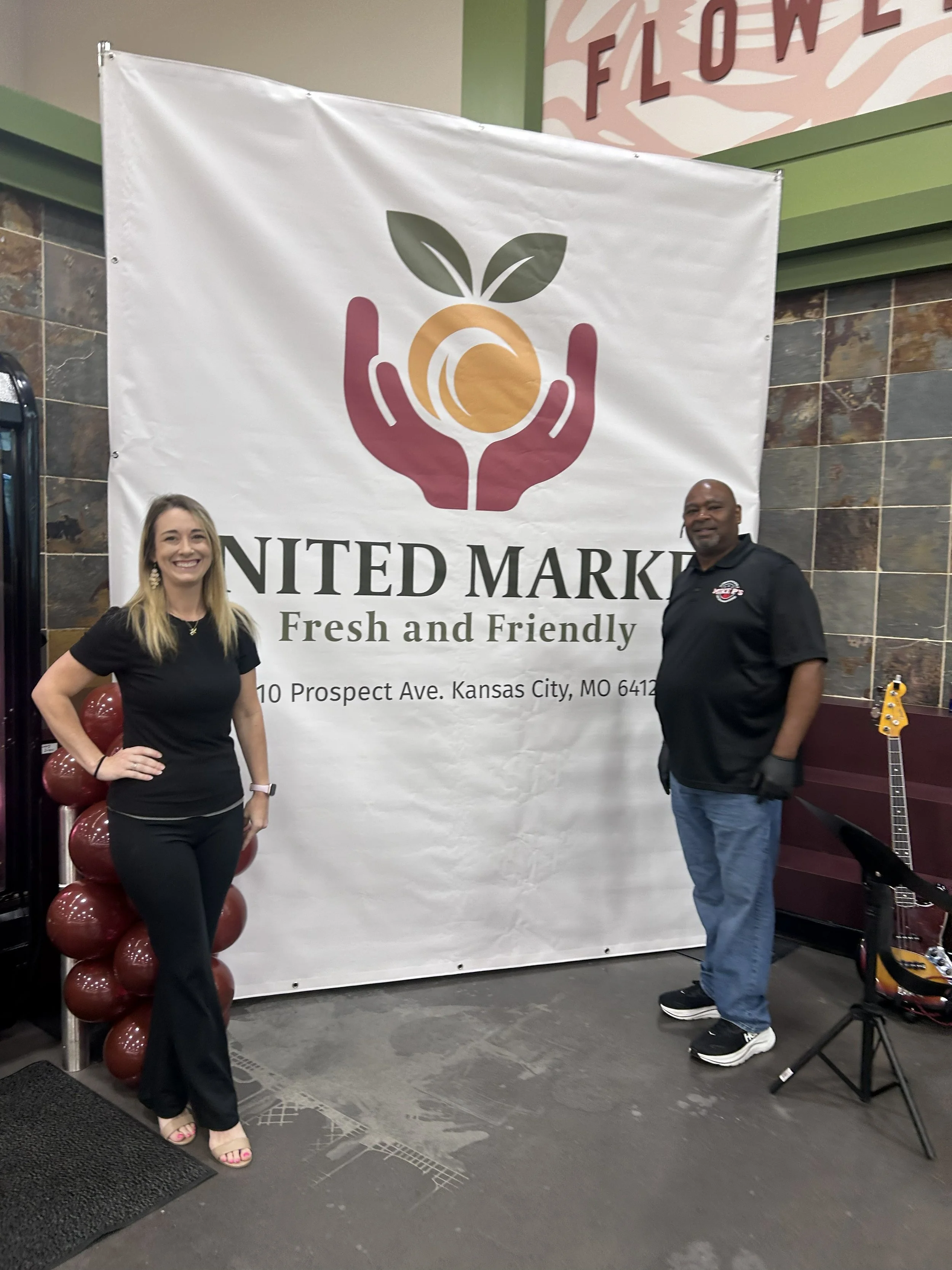 Two people standing in front of a banner with a logo of a hand holding an orange with leaves, and text that reads "United Market - Fresh and Friendly" at a store or event. The woman on the left is smiling, wearing a black shirt and black pants, with long blonde hair. The man on the right is wearing a black polo shirt and jeans, with a bald head and a goatee, standing near a guitar and a music stand.