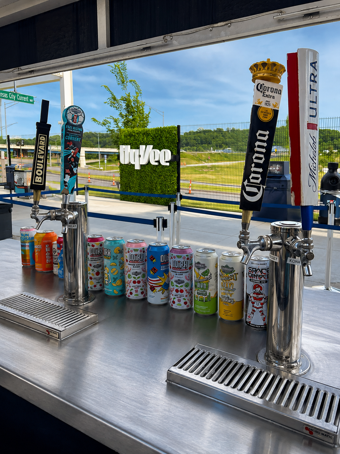 A row of colorful canned beverages displayed on a stainless steel bar with beer taps labeled 'BOULEVARD', 'Corona Extra', and 'Michelob Ultra'.