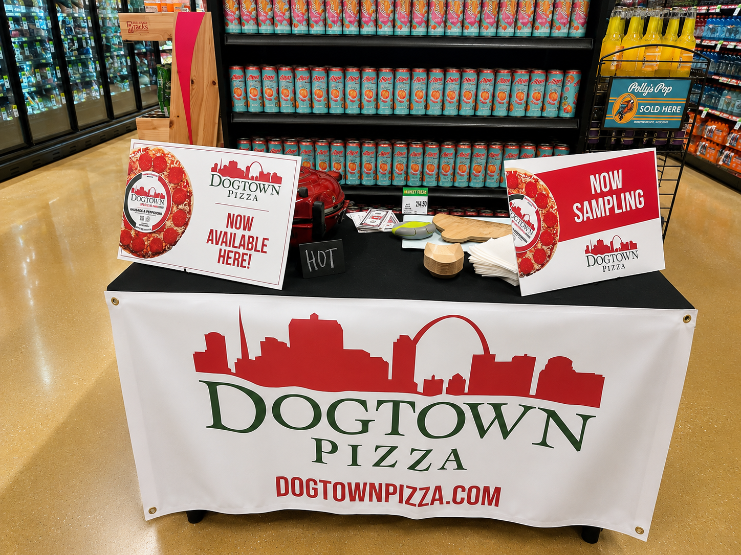 A promotional display for Dogtown Pizza set up on a table in a grocery store, with signs indicating availability and sampling, and shelves stocked with canned drinks and bottled beverages in the background.