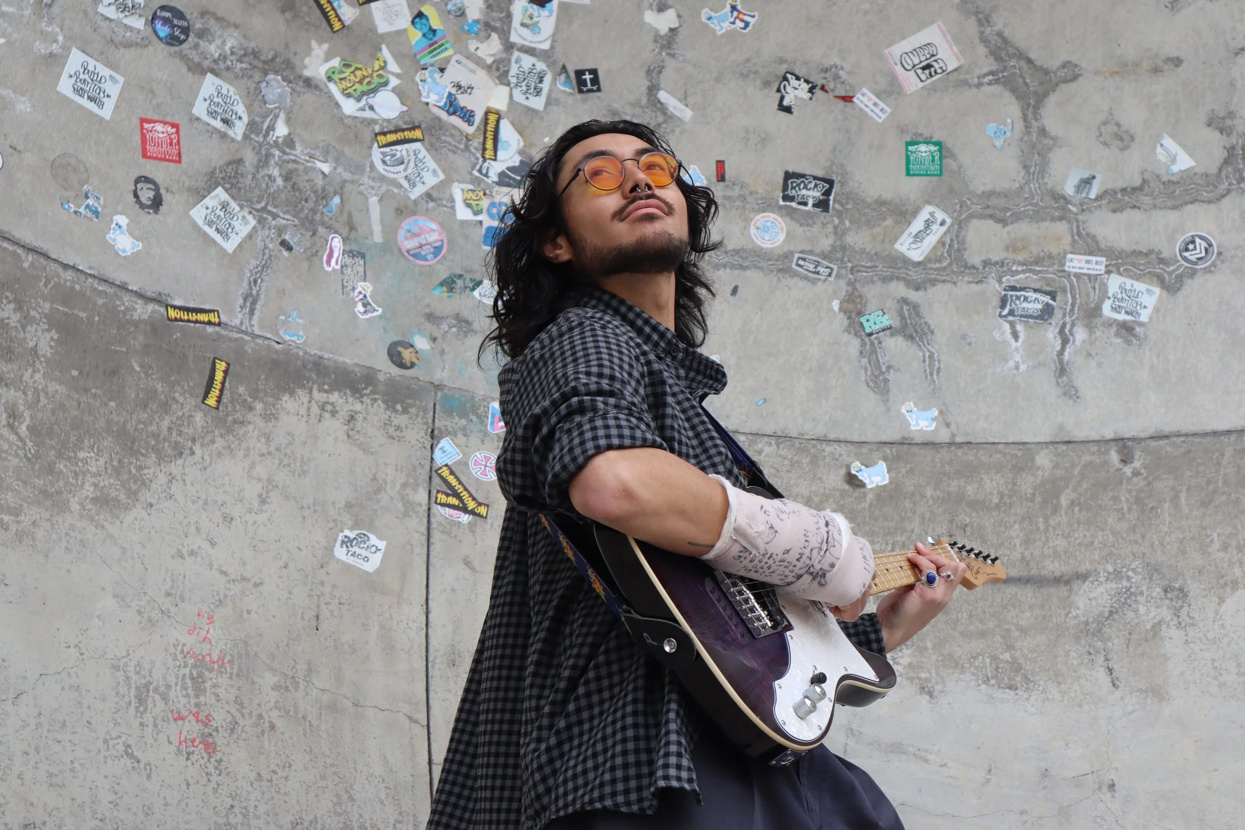young man at the skate park with long hair orange glasses and a guitar