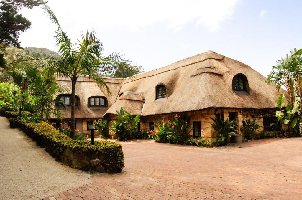 A thatched-roof house surrounded by lush greenery and tropical plants, with a brick driveway and a mountain in the background.