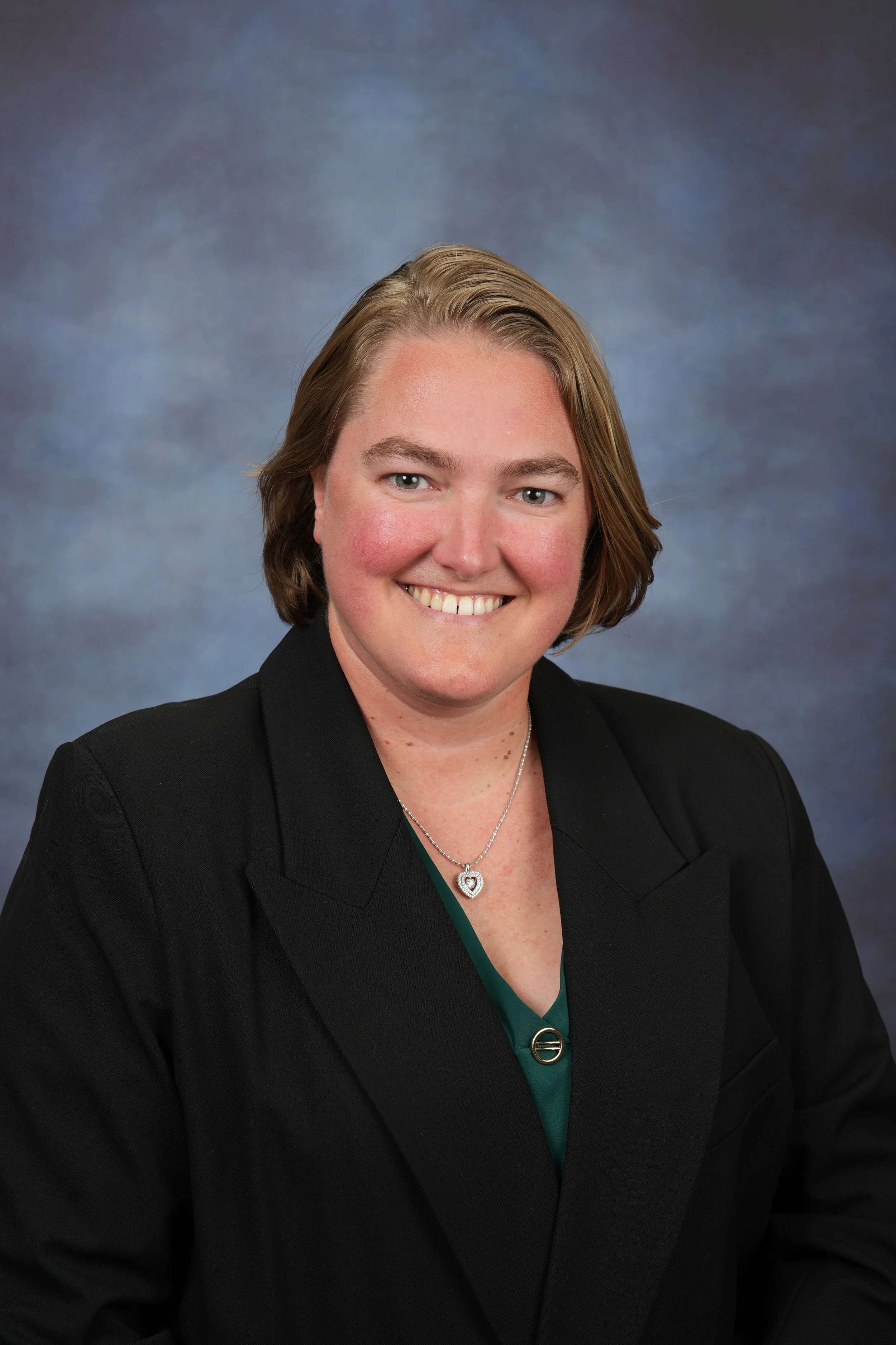 Portrait of a smiling woman with short blonde hair, wearing a black blazer and a silver necklace with a heart-shaped pendant, against a gray and blue mottled background.