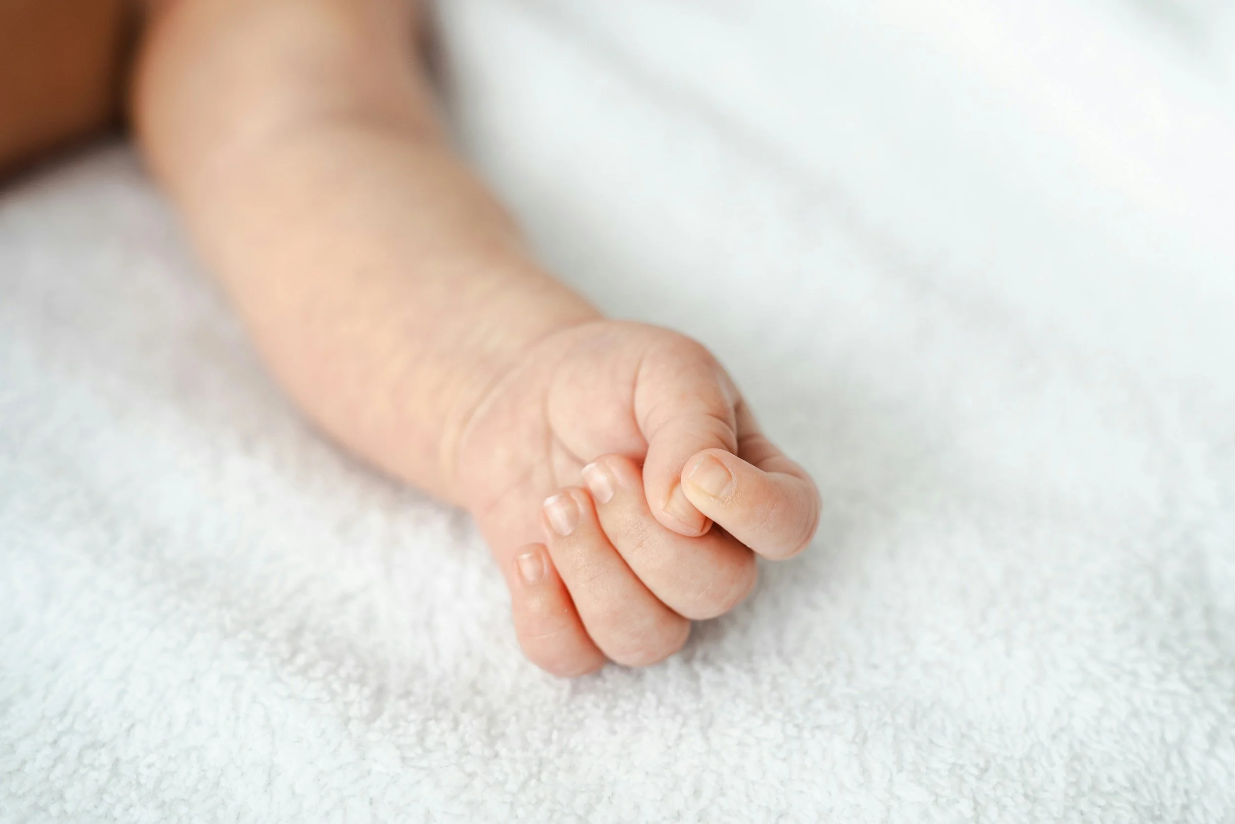 Close-up of an adult hand gently holding a baby's hand, both resting on a soft, white surface.