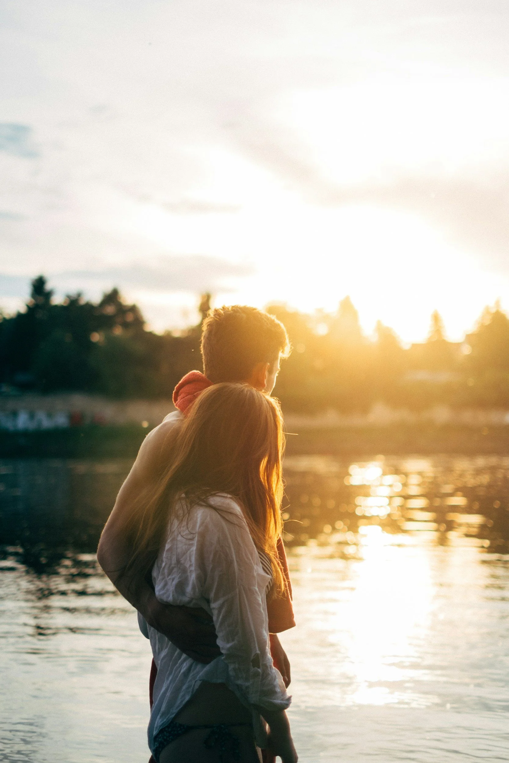 Two children, a boy and a girl, standing by a body of water at sunset, facing away from the camera.