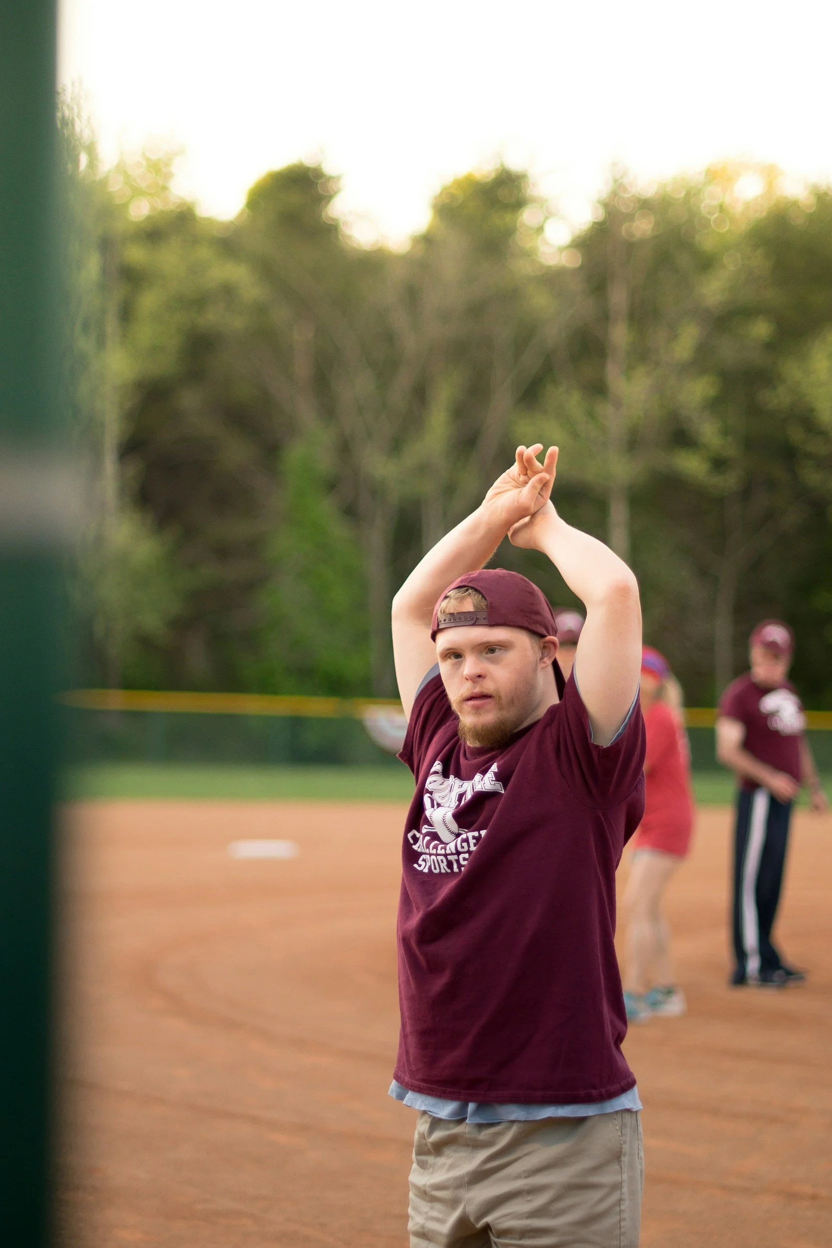 A young man with a red cap backwards and a maroon T-shirt with a sports logo, standing on a baseball field with his arms raised, participating in a game or practice. Other people are in the background, and there are trees and a bright sky.