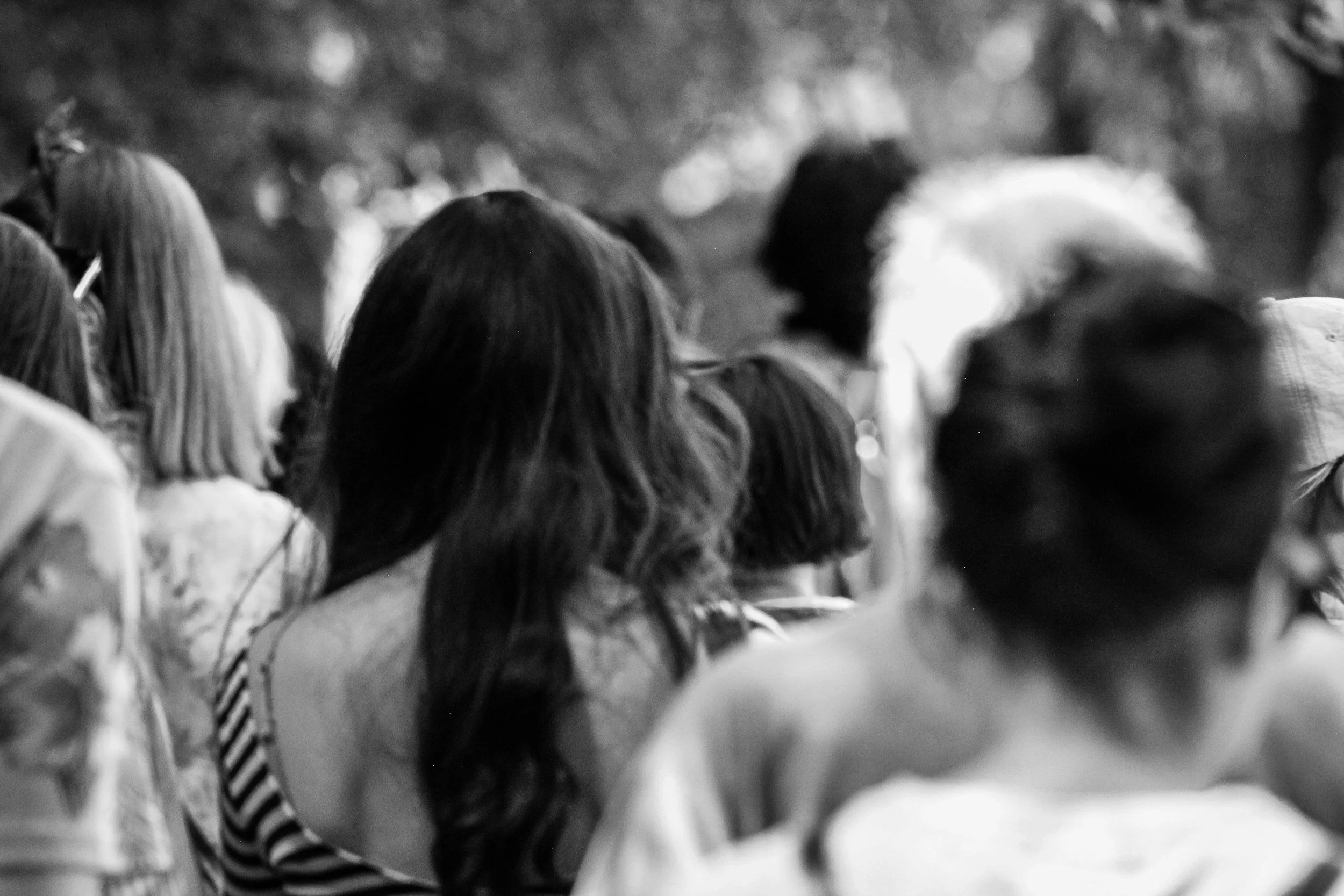 Black and white photo of a crowd of people, mostly women with long hair, facing away from the camera in an outdoor setting.