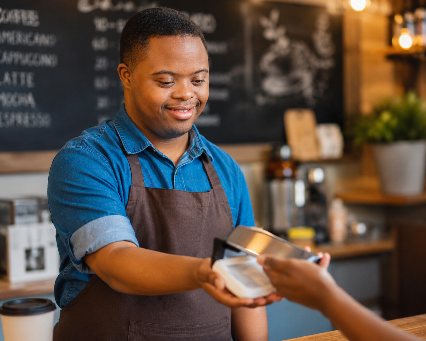 A barista in a blue denim shirt and brown apron is smiling as they take a payment card from a customer in a coffee shop.