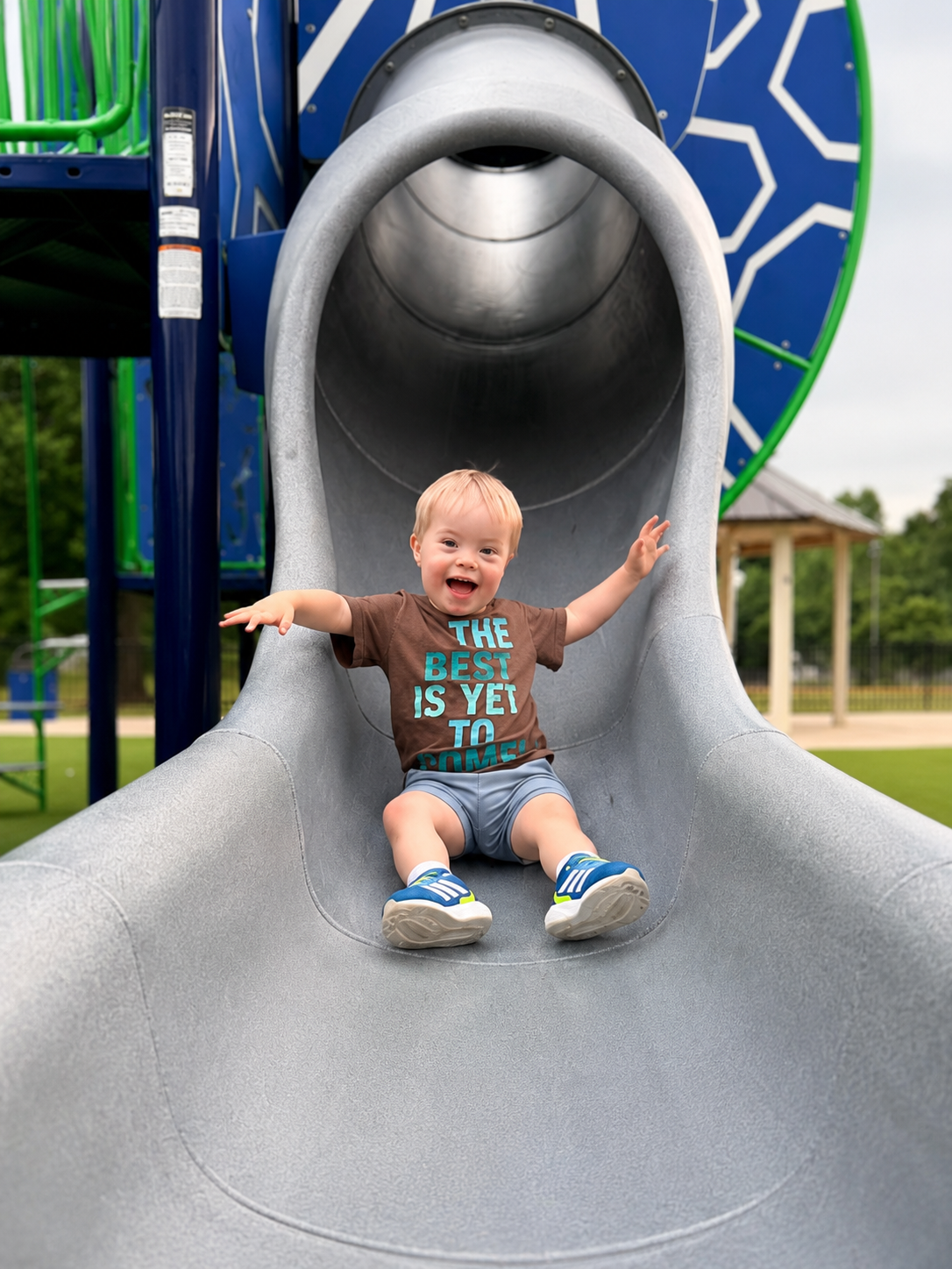 A happy young boy sitting at the bottom of a playground slide with a tunnel at a park, smiling and raising his arms.