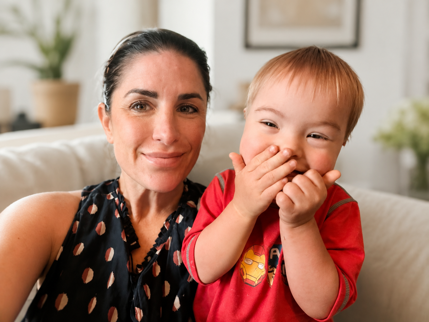 A woman and a young boy sitting on a sofa, smiling and laughing. The woman has dark hair and is wearing a sleeveless top, while the boy has red hair and is wearing a red shirt with a superhero logo.