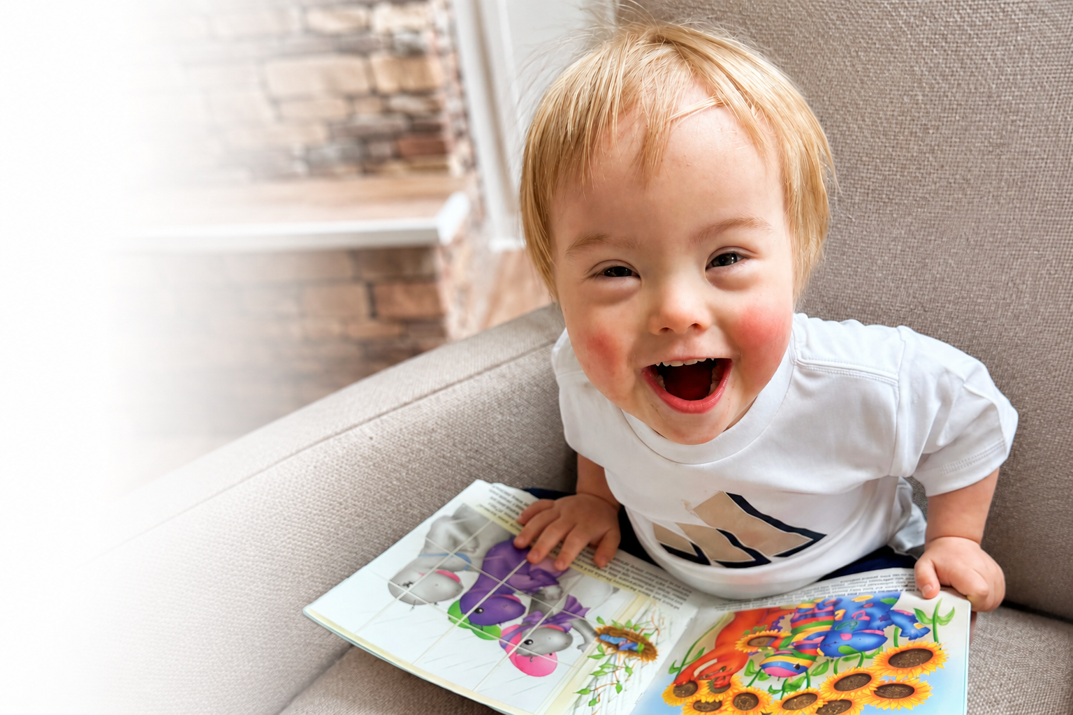 A happy young boy with blond hair sitting on a beige couch, smiling widely with mouth open, looking up at the camera, with an open children's book about animals and sunflowers in front of him.