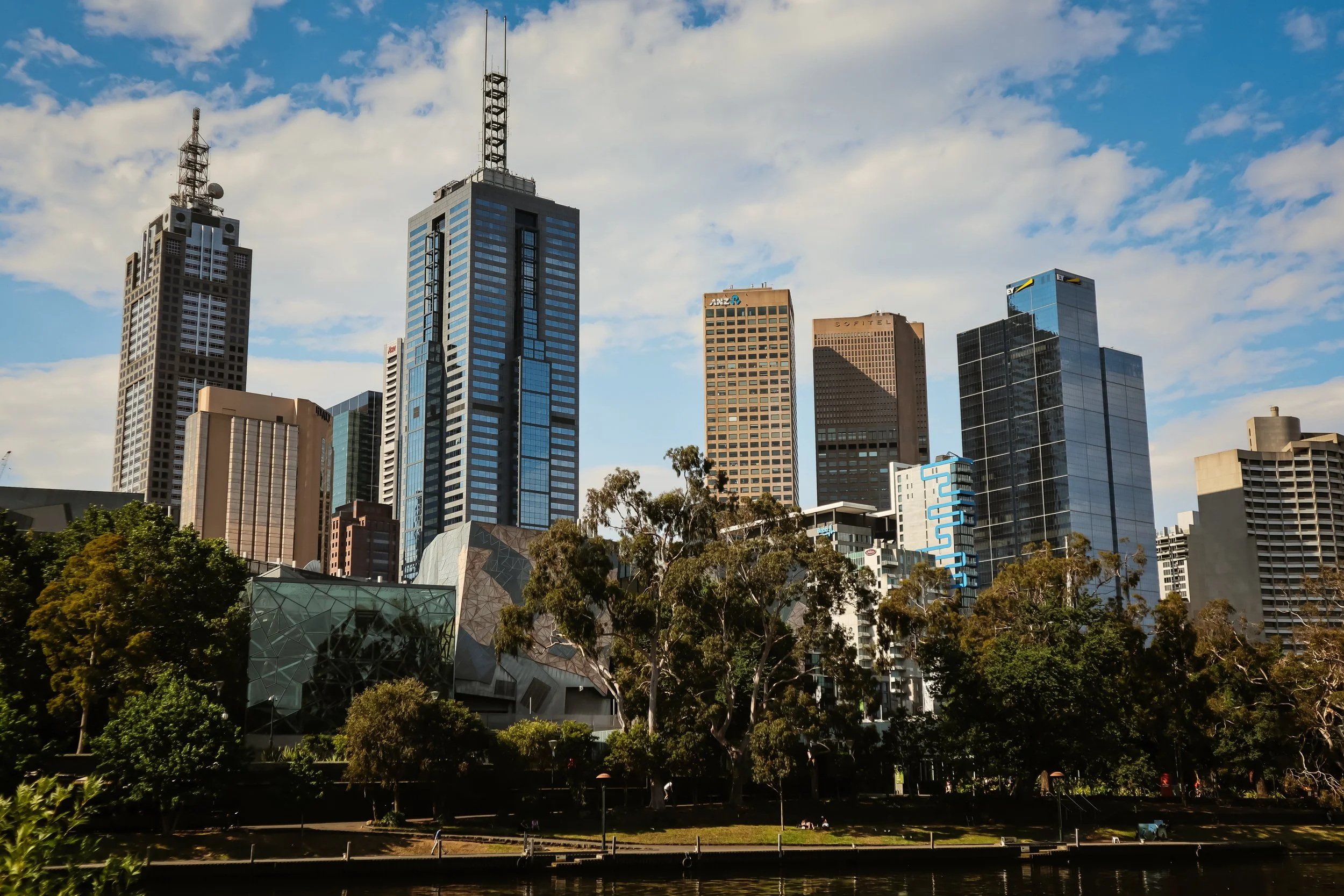 City skyline with tall modern buildings and skyscrapers, some with glass exteriors, set against a partly cloudy sky, with trees and a park in the foreground.