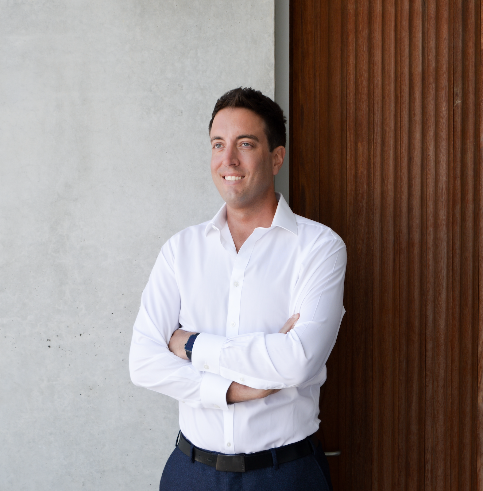 A man with short dark hair wearing a white dress shirt, dark blue pants, and a watch, standing with arms crossed and smiling, leaning against a wall with a wooden panel on one side.