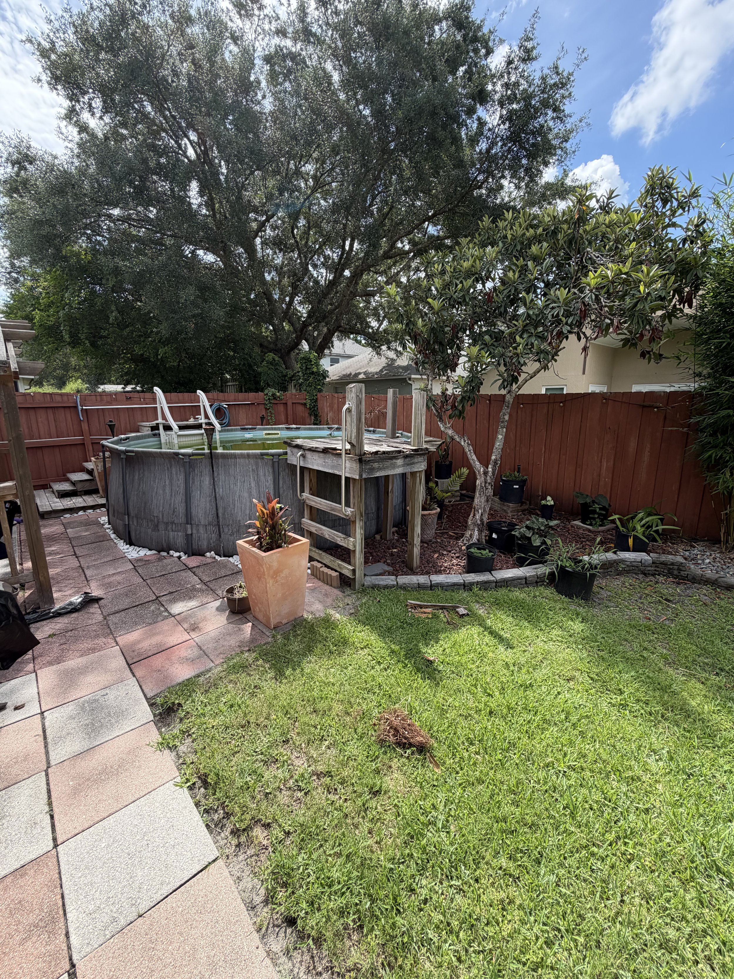 A backyard with a pond surrounded by potted plants, a tree, a wooden fence, and a patio with paving stones, under a partly cloudy sky.