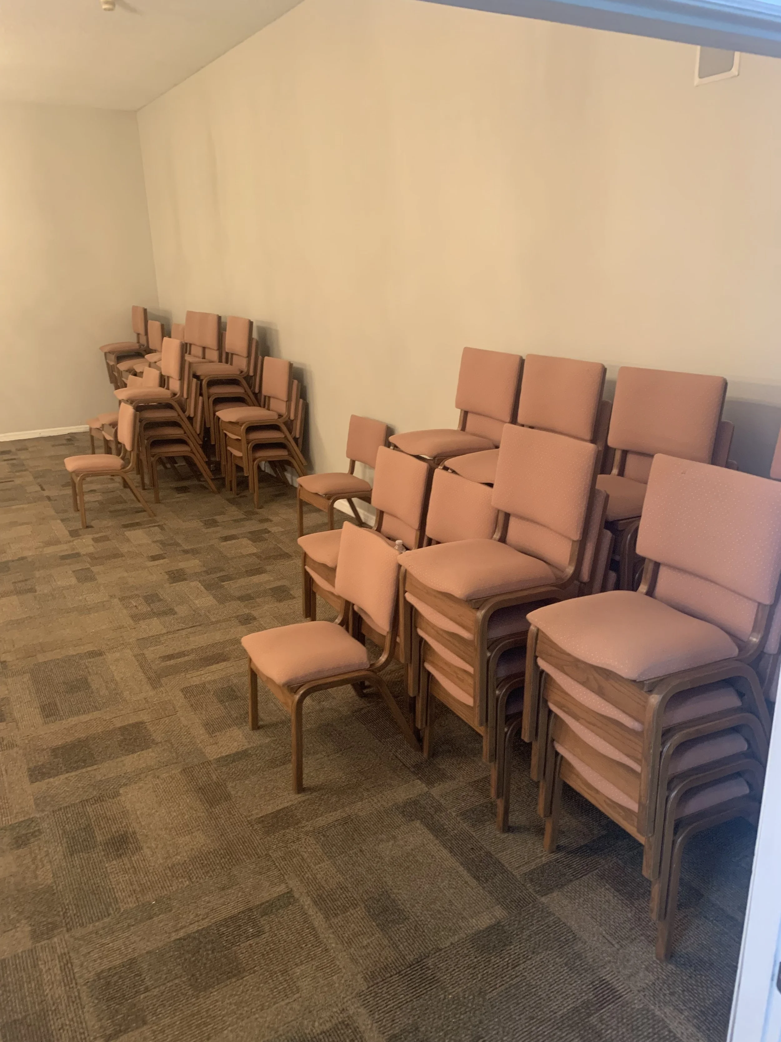 Stacked beige cushioned chairs inside a room with a beige and brown checkered carpet and plain beige walls.