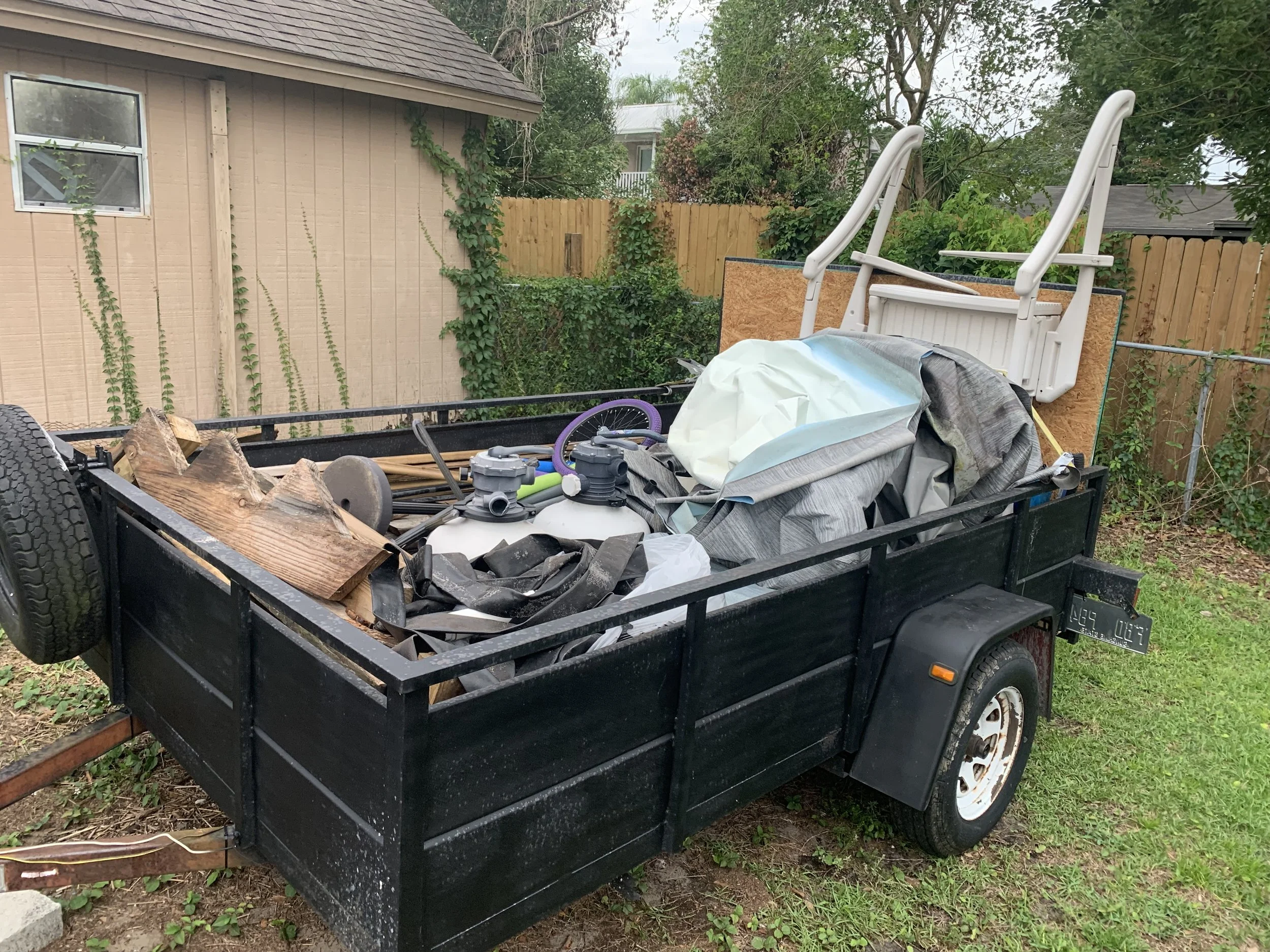 A black utility trailer filled with logs, gardening tools, and tarp, parked on grass in a backyard with a wooden house and a wooden fence, surrounded by trees and shrubs.