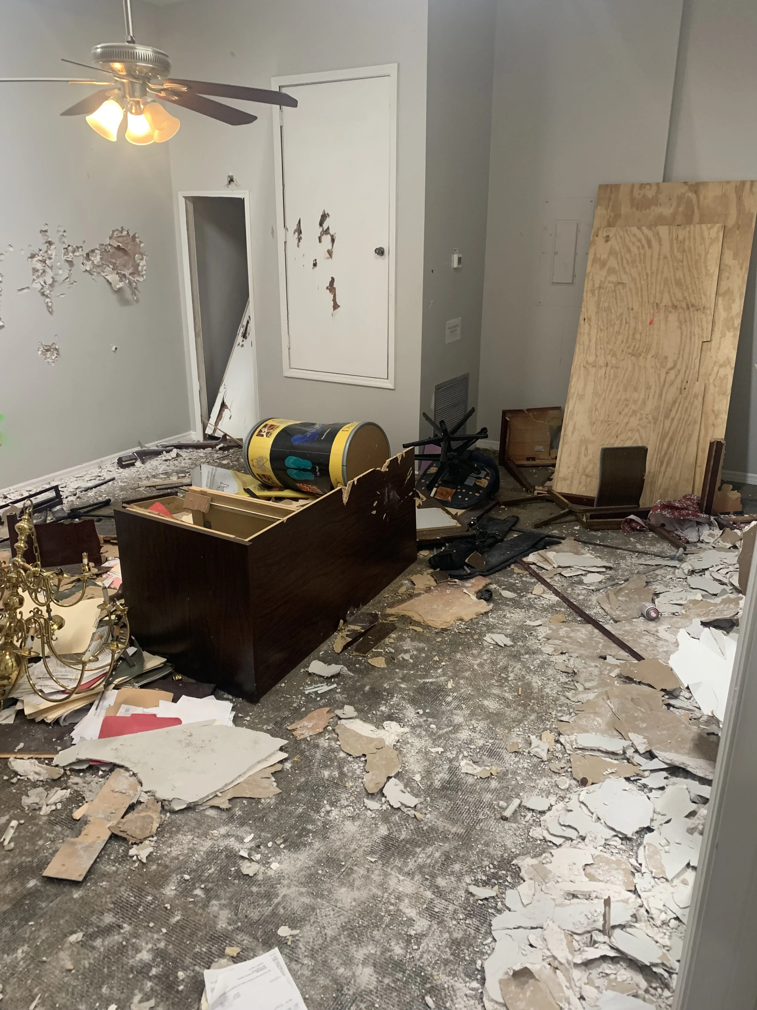 A room in disarray with fallen ceiling debris, broken drywall, and scattered papers. A ceiling fan with four lights is turned on. There is a damaged wall with peeling paint, and various furniture and objects are overturned or damaged.