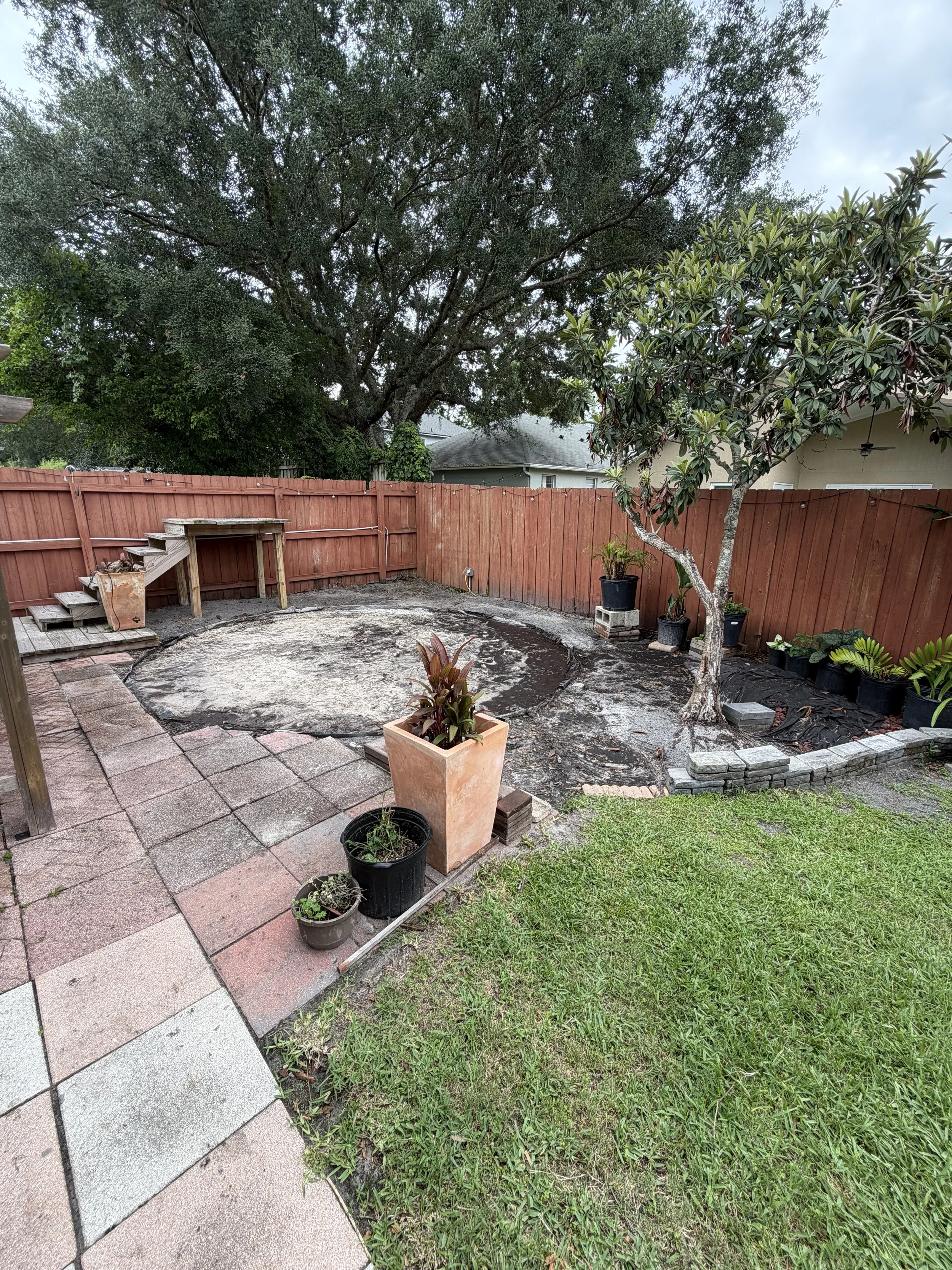 Backyard with a wooden fence, a large tree, potted plants, a small raised garden bed, and a tiled patio area.
