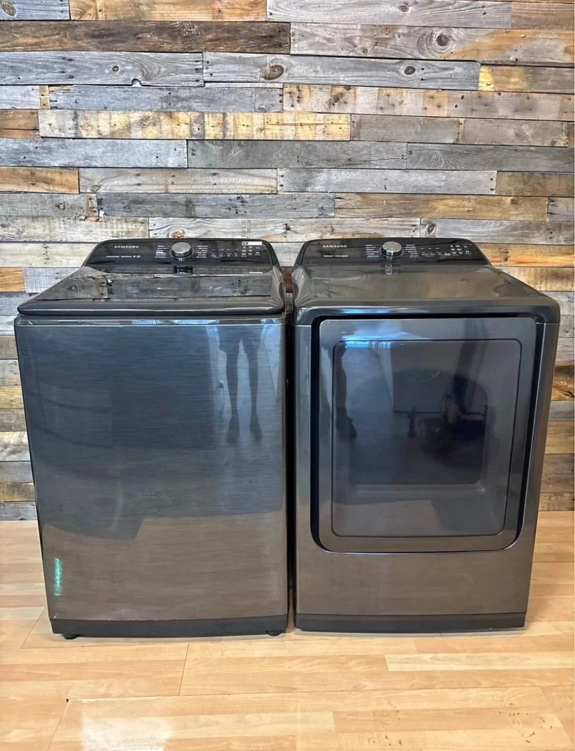 A pair of black and silver washing machines, one with a top-loading design and the other with a front-loading door, set against a rustic wooden wall.