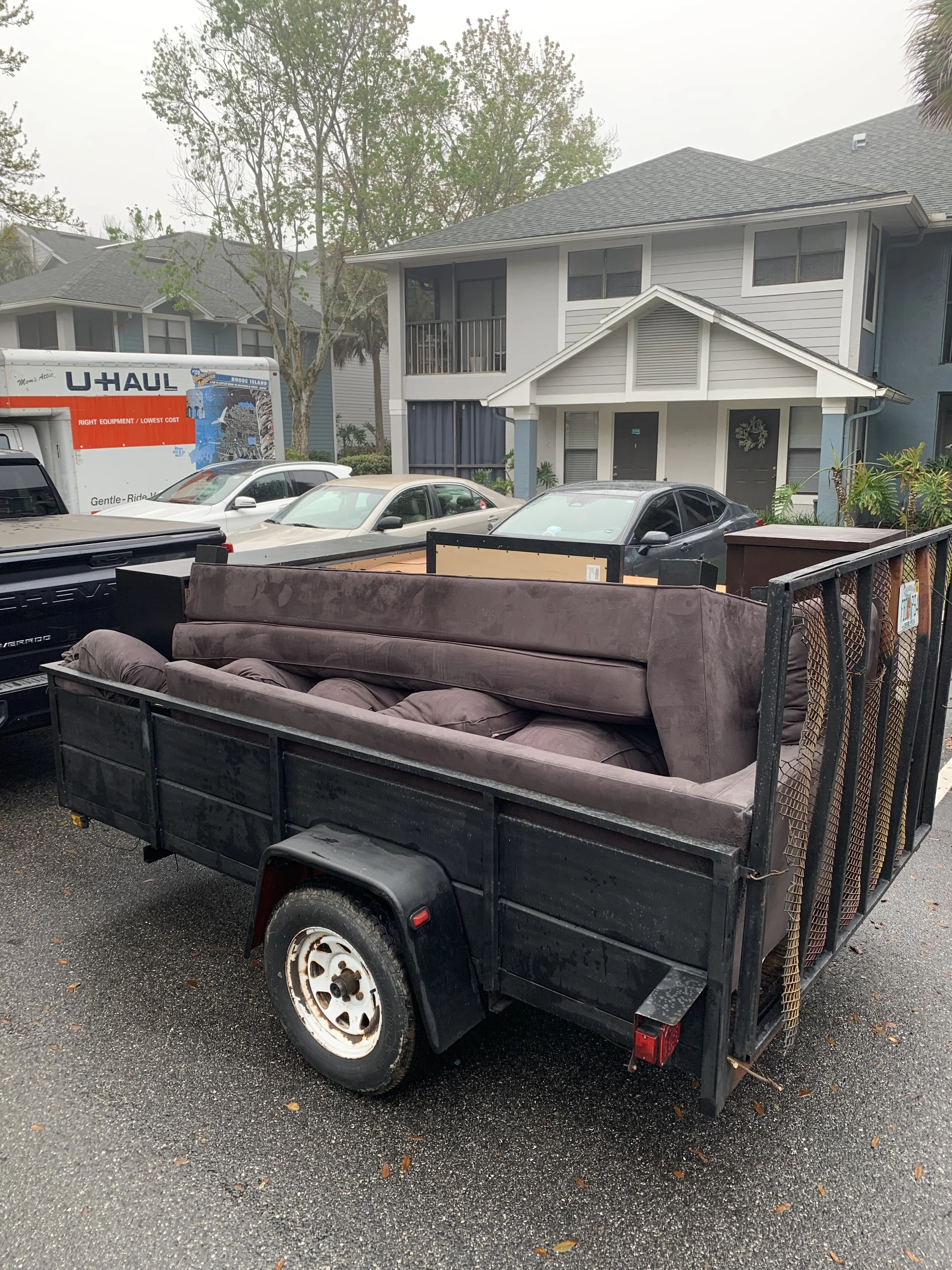A small black trailer carrying a brown sofa with cushions parked on a wet driveway in front of a blue and white two-story house with several cars and trees in the background.