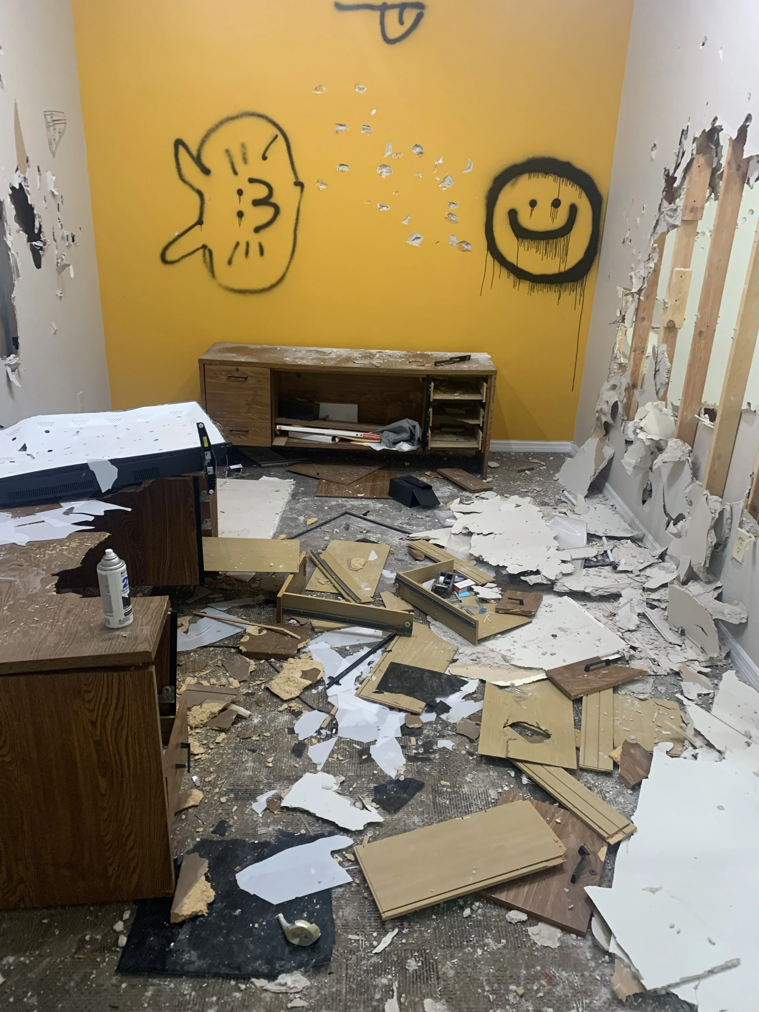 A room with a yellow accent wall featuring graffiti drawings of a smiling face and a character with pointed ears, and a broken wall with torn drywall. The floor is filled with debris, broken drywall, and scattered materials. There is a wooden cabinet against the yellow wall, a disorganized table with torn paper and a marker, and various broken pieces of wood and drywall strewn across the room.