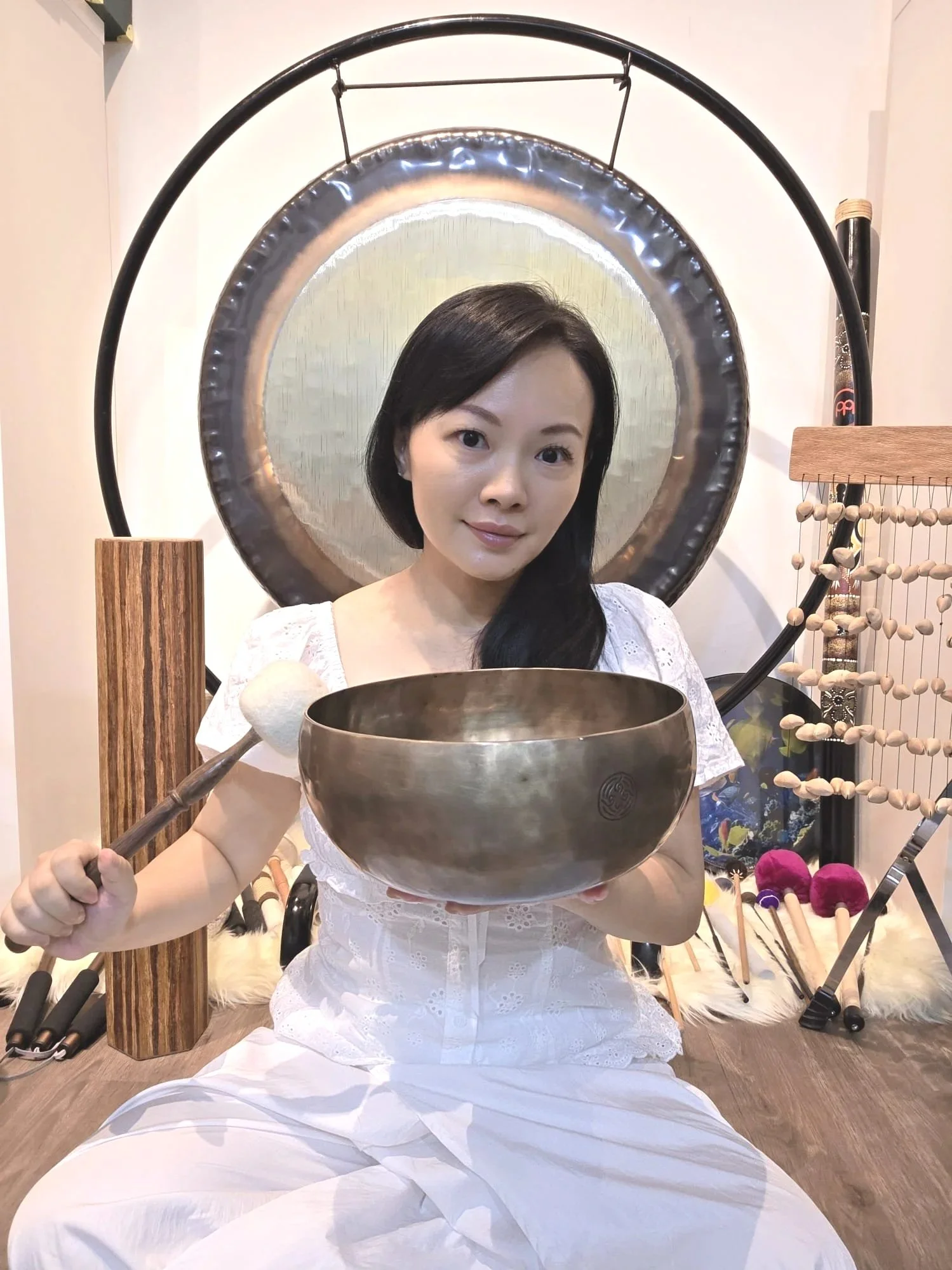 Woman dressed in white, sitting on the floor, holding a singing bowl and a mallet, with musical instruments and decorative items around her, a large gong hanging in the background.