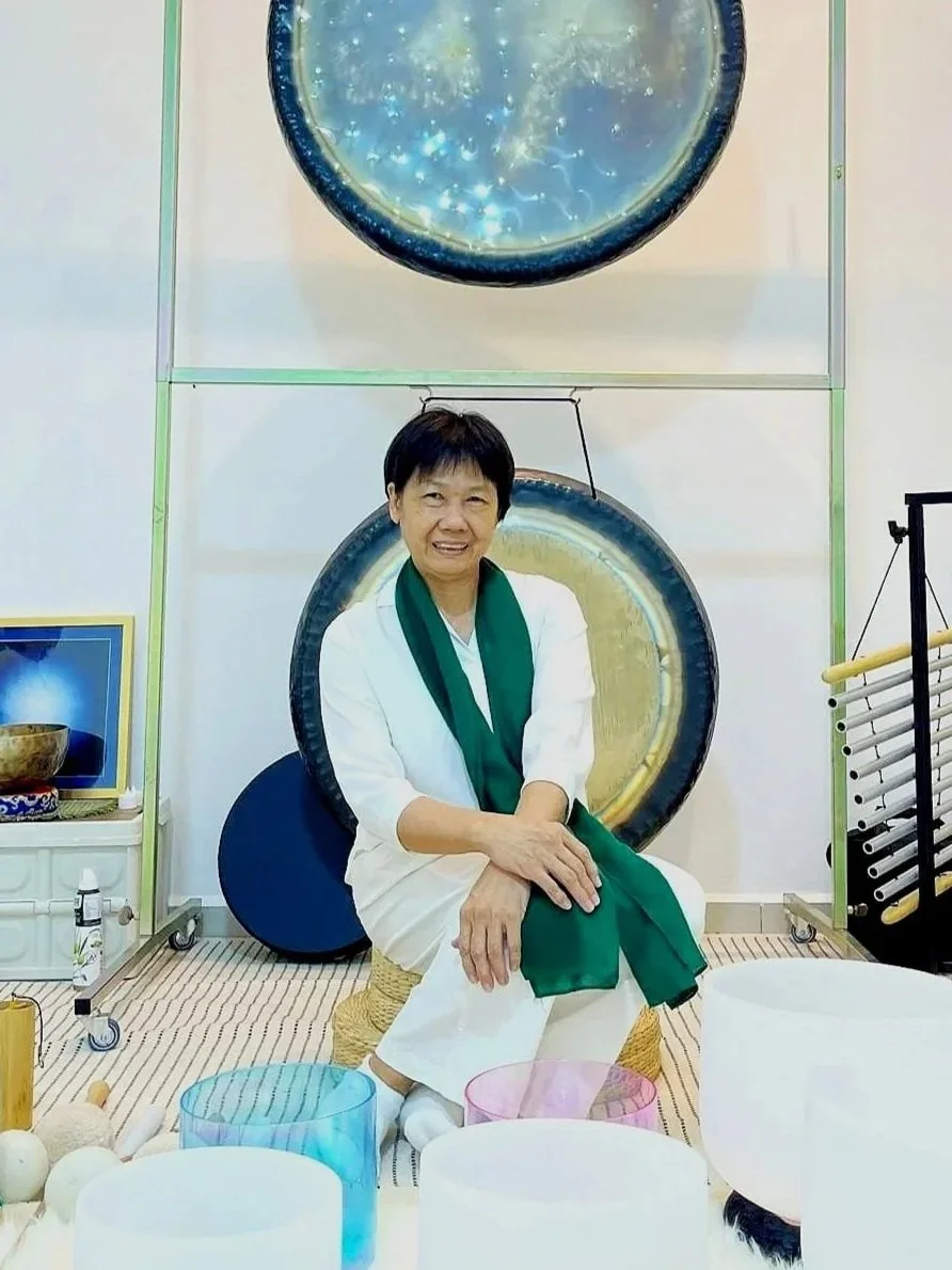 A woman sitting on a small stool with a drum in front of her, surrounded by singing bowls and musical instruments, with two gongs hanging on the wall behind her.