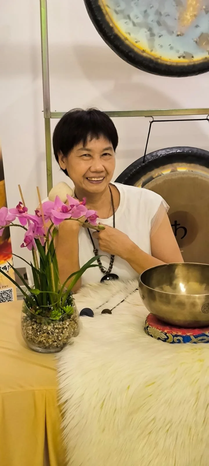 A woman sitting on a small stool with a drum in front of her, surrounded by singing bowls and musical instruments, with two gongs hanging on the wall behind her.