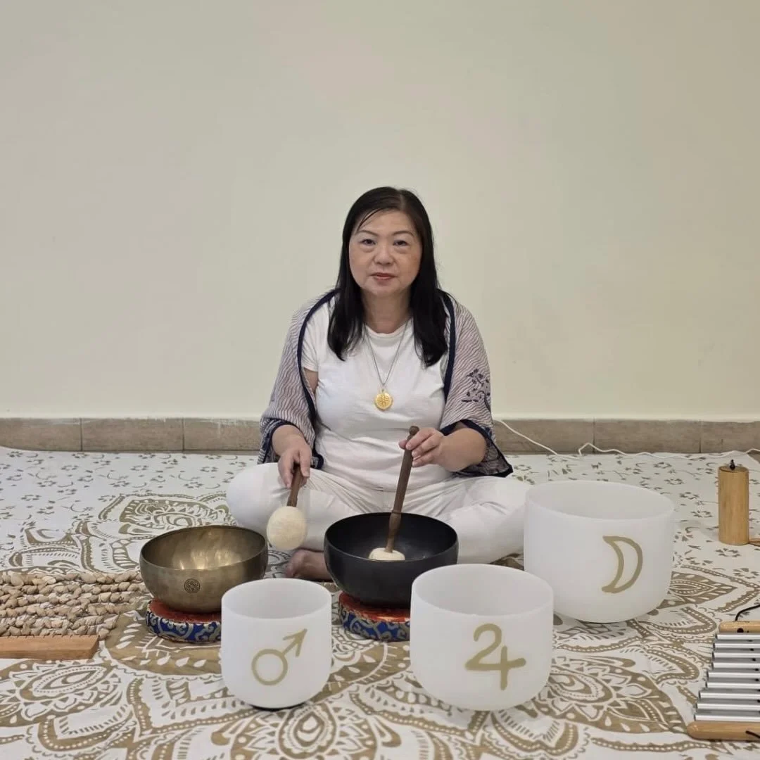 A woman with black hair sitting cross-legged on a white and gold patterned cloth, surrounded by crystal singing bowls, a Tibetan bell, and various other instruments, holding mallets, in a minimalistic room with a plain beige wall.