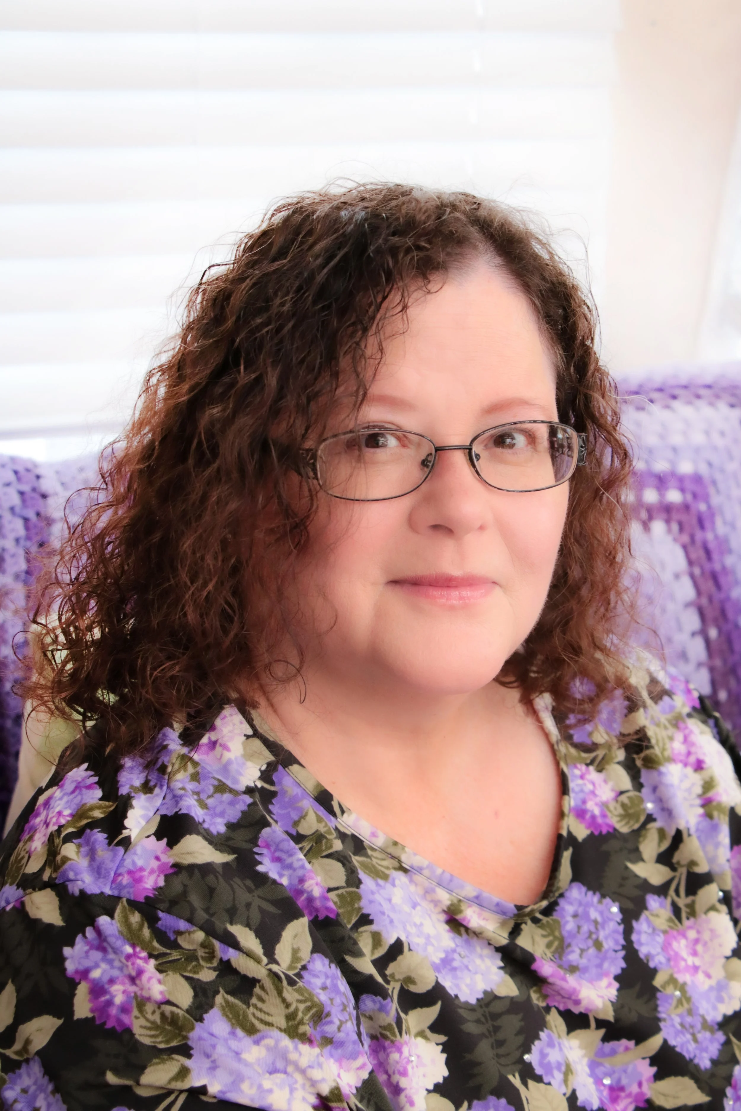 A woman with curly brown hair, wearing glasses and a floral blouse with purple flowers, sitting on a purple crocheted blanket.