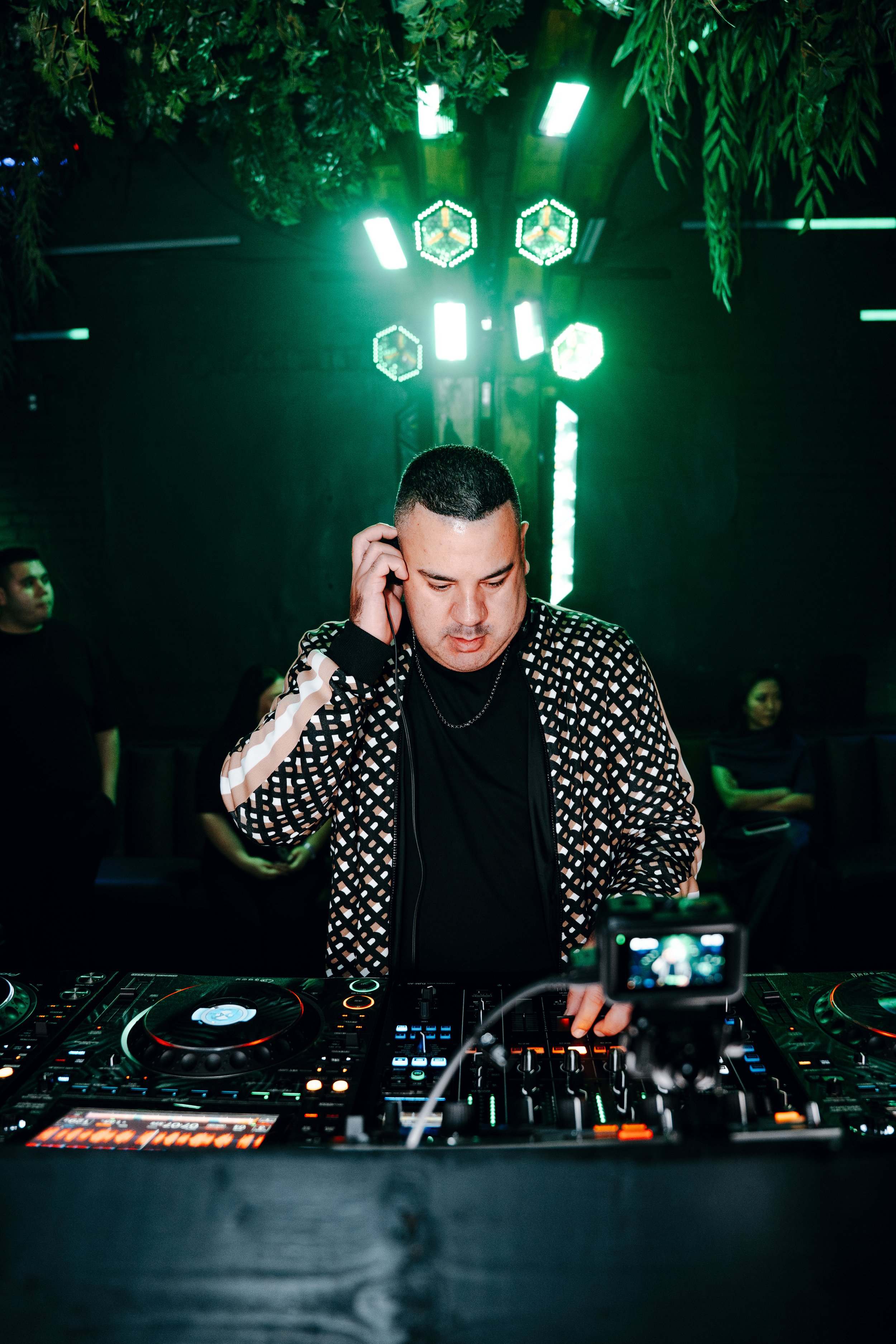 A DJ wearing a black and white checkered jacket and black shirt, using DJ equipment at an event with green lighting and tropical foliage, while a person in the background observes.