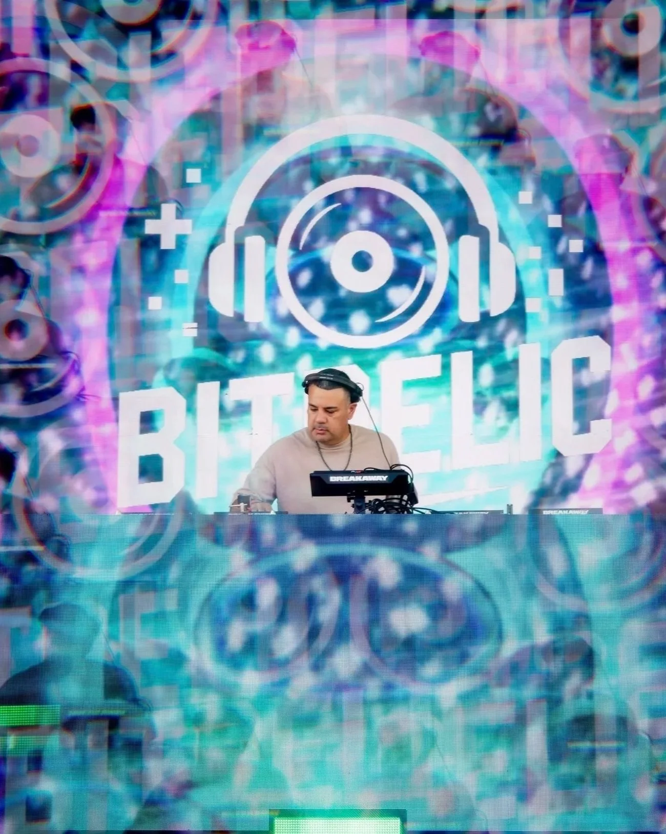 A DJ performing on stage with a digital backdrop displaying the words 'Bitgenic' and a headphone logo, surrounded by vibrant, colorful visuals.