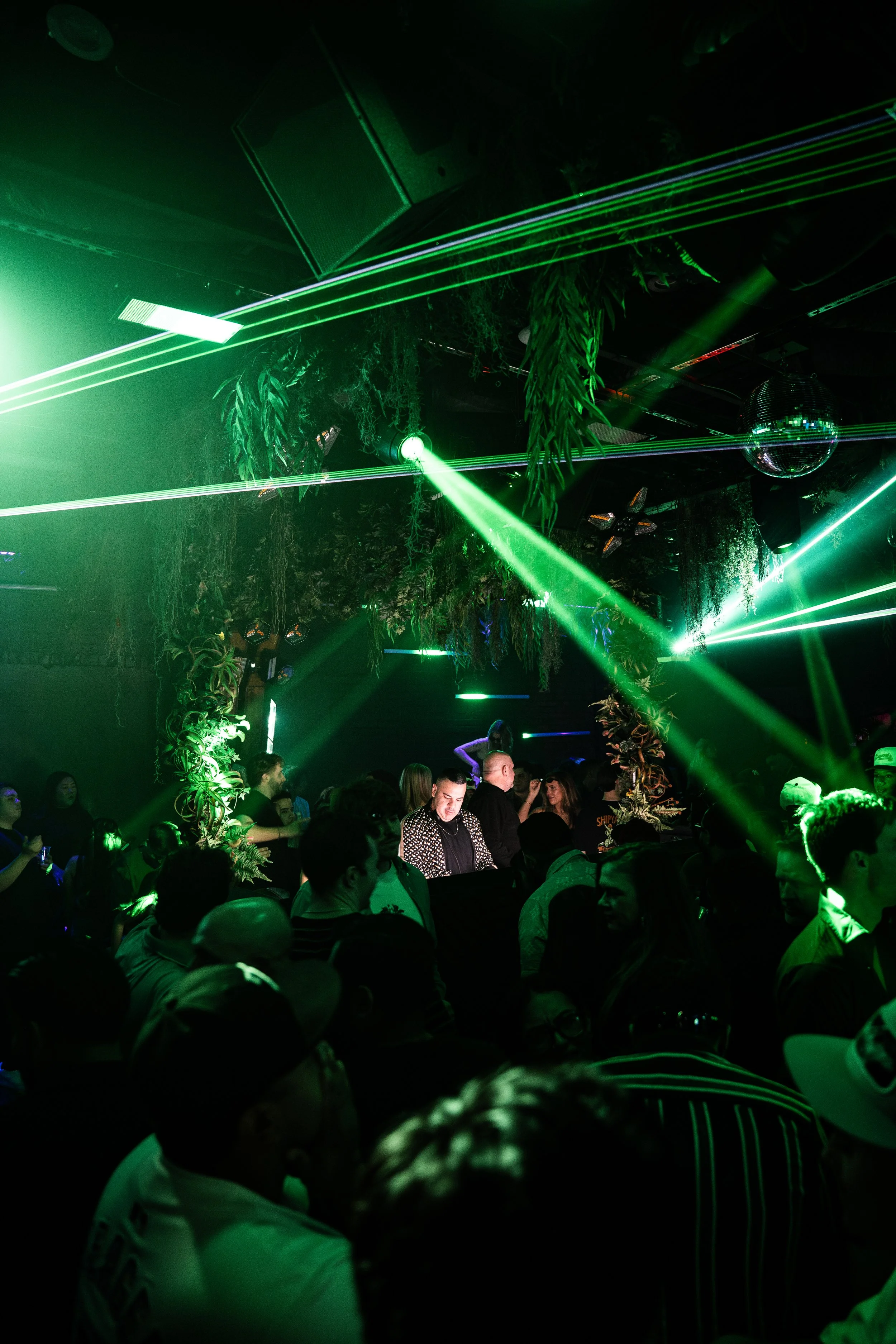 A crowded nightclub scene with green laser lights, hanging plants, and people dancing and socializing.