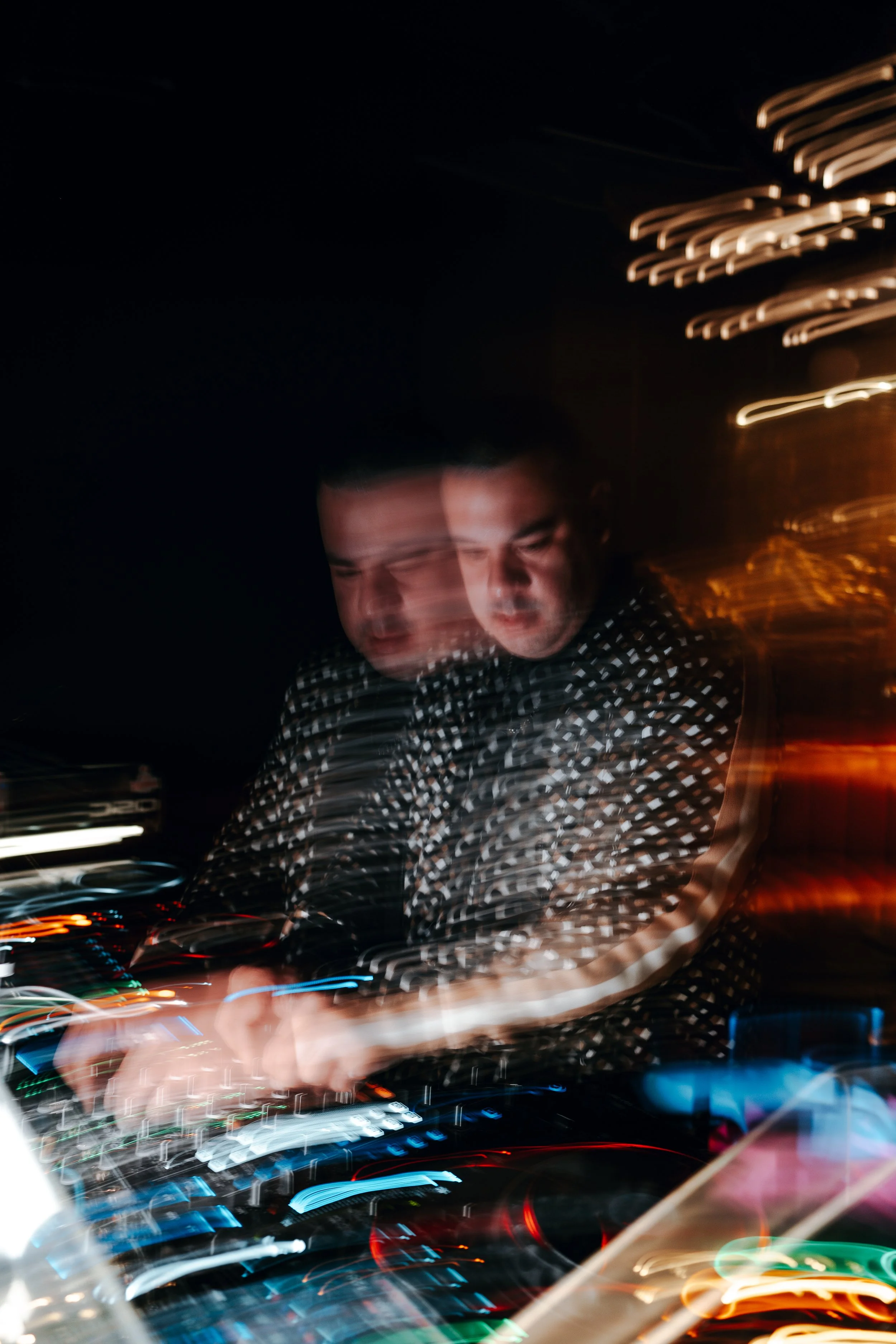 A person with short dark hair wearing a black and white checkered shirt, focused on a DJ mixer with colorful lights at night, with a blurred background of a building with illuminated windows.