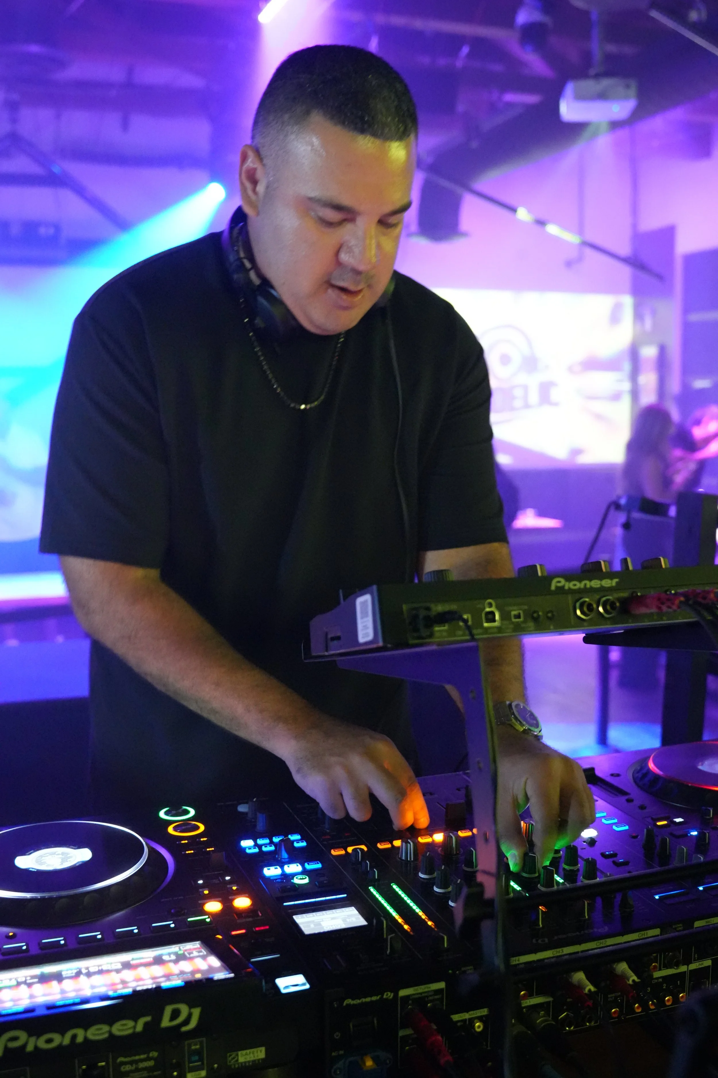 A DJ wearing a black t-shirt and a watch is performing in a club with colorful lights and a DJ controller in front of him.