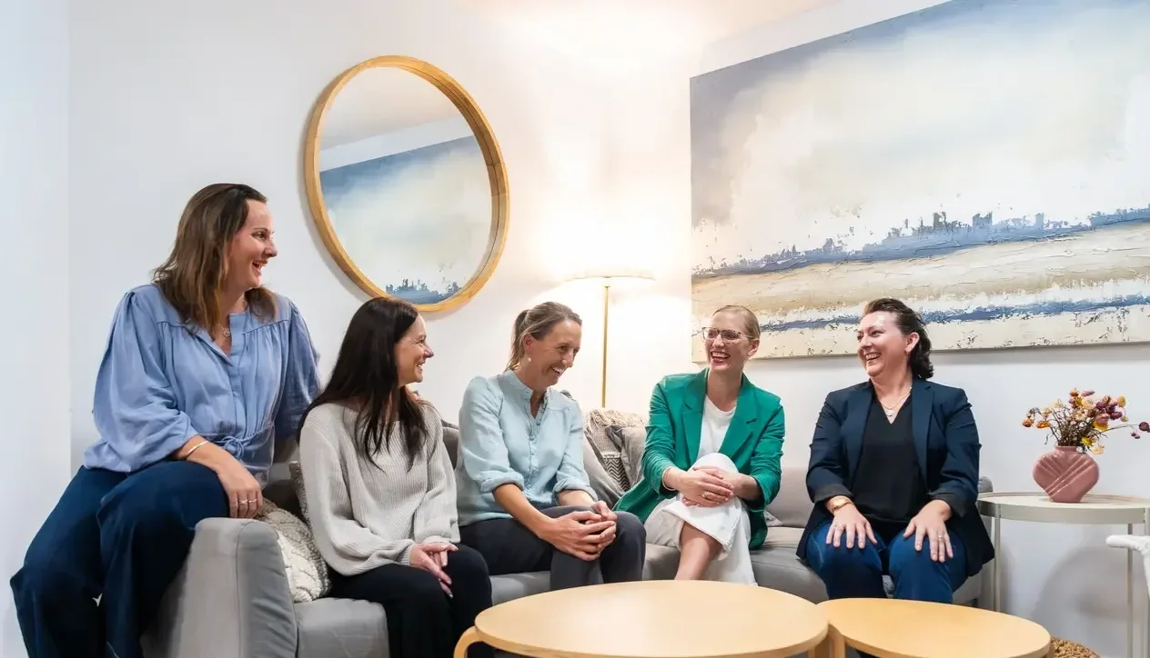 Five women sitting and standing on a gray couch, engaging in conversation and laughing in a well-lit living room with artwork and a table with a pink vase of flowers.