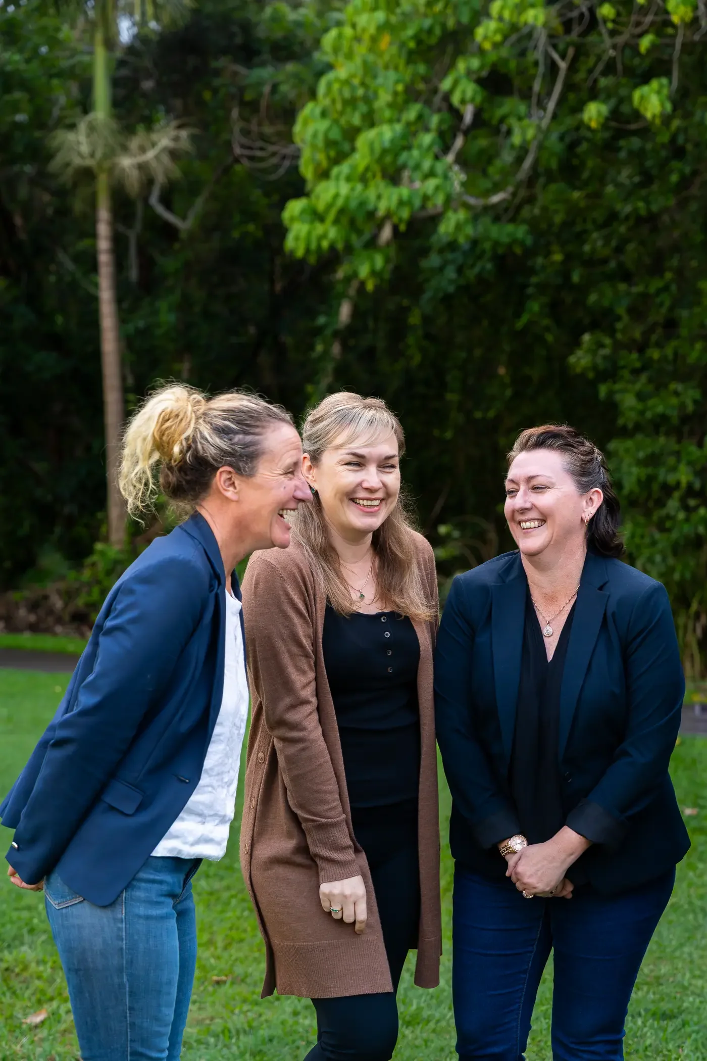 Three women standing outdoors on a lawn, smiling and engaging in conversation, with trees and greenery in the background.