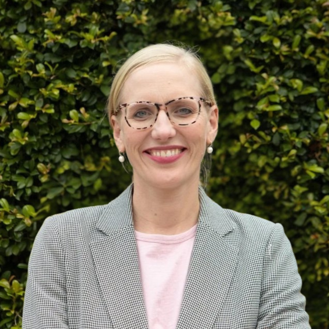 A woman with blonde hair and glasses smiling in front of a green leafy background.