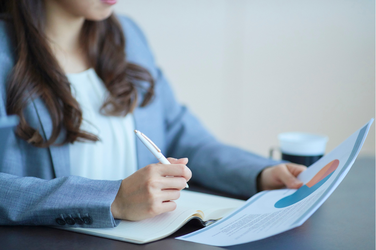 A woman sitting at a table, holding a pen and reviewing a document with pie charts, with a cup in the background.