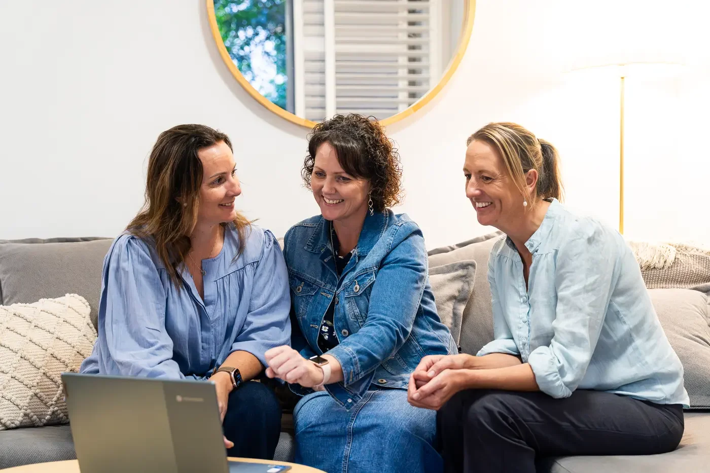 Three women talking happily on a sofa in a living room with a round mirror and a lamp behind them.