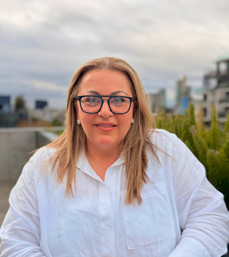 Woman wearing glasses smiling, outdoor urban setting.