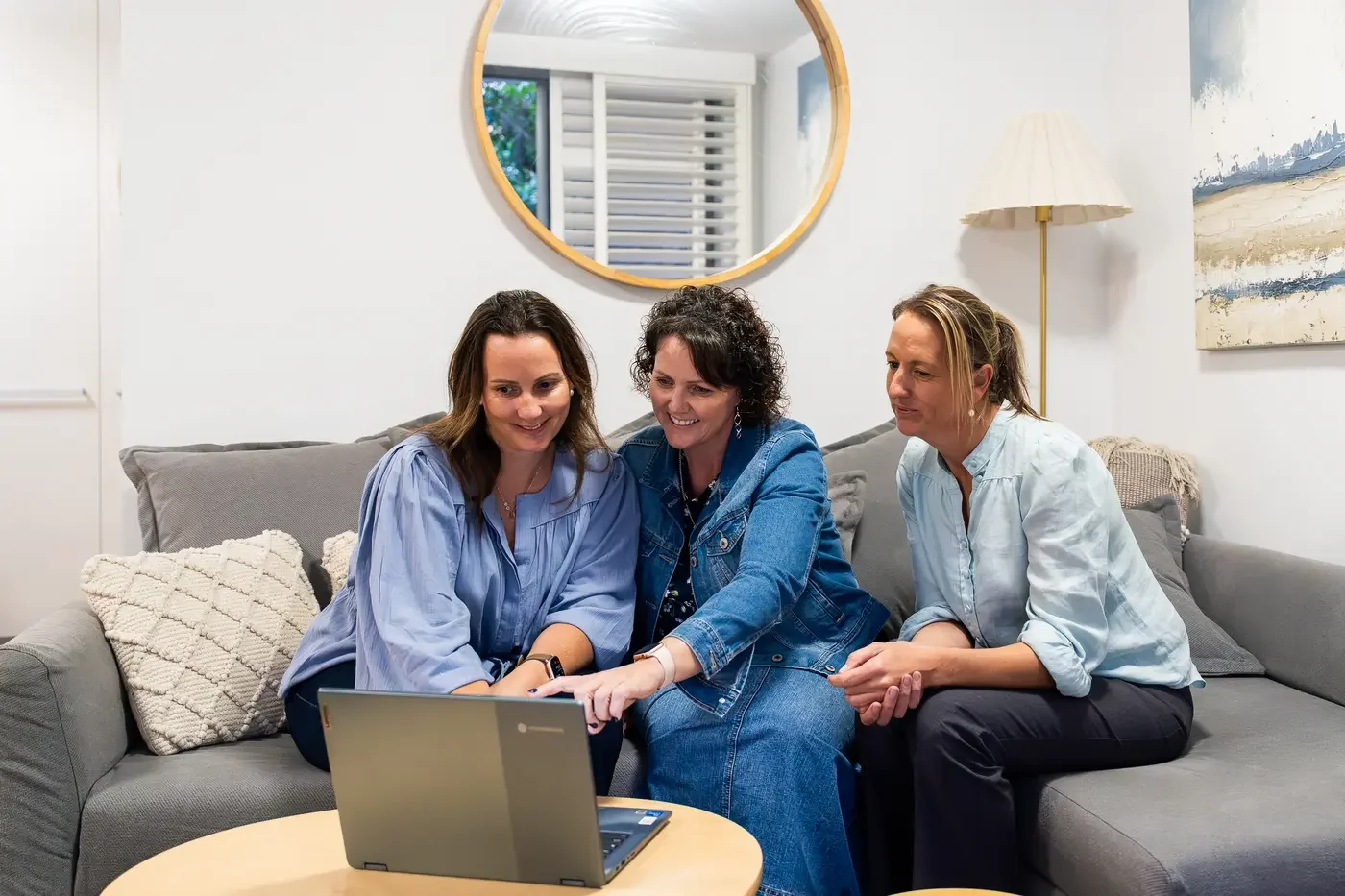Three women sitting on a gray couch, looking at a laptop, smiling, in a cozy living room with a mirror, lamp, and wall art.