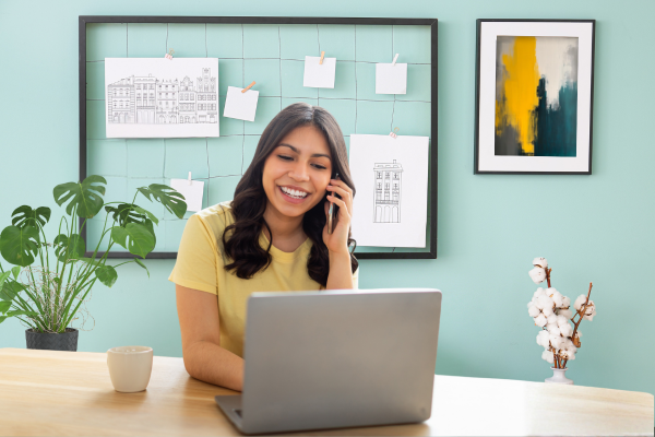 Employee engaging in EAP counselling on phone. She's wearing a yellow T-shirt, is smiling and looking at her laptop. on the background there's a plant, some flowers, some pictures pegged to a frame, and some framed art. The back wall is teal.