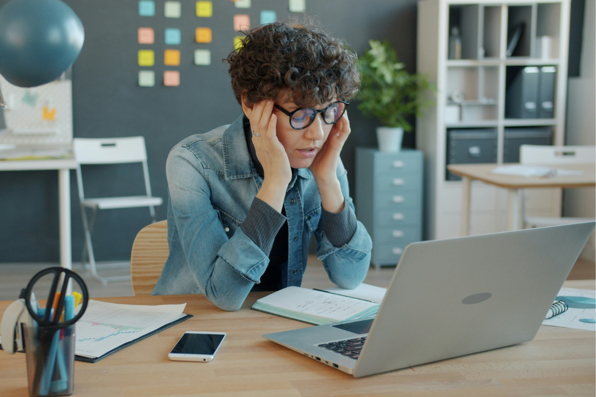 A woman with curly hair and glasses sitting at a desk, holding her head in frustration while looking at her open laptop. The desk has papers, a smartphone, and office supplies, with a modern office background.