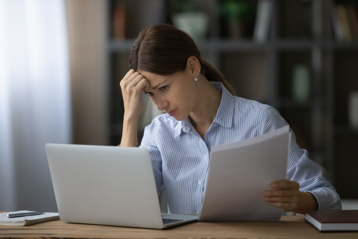 Stressed woman working at desk with laptop, papers, and books, in a room with bookshelves.