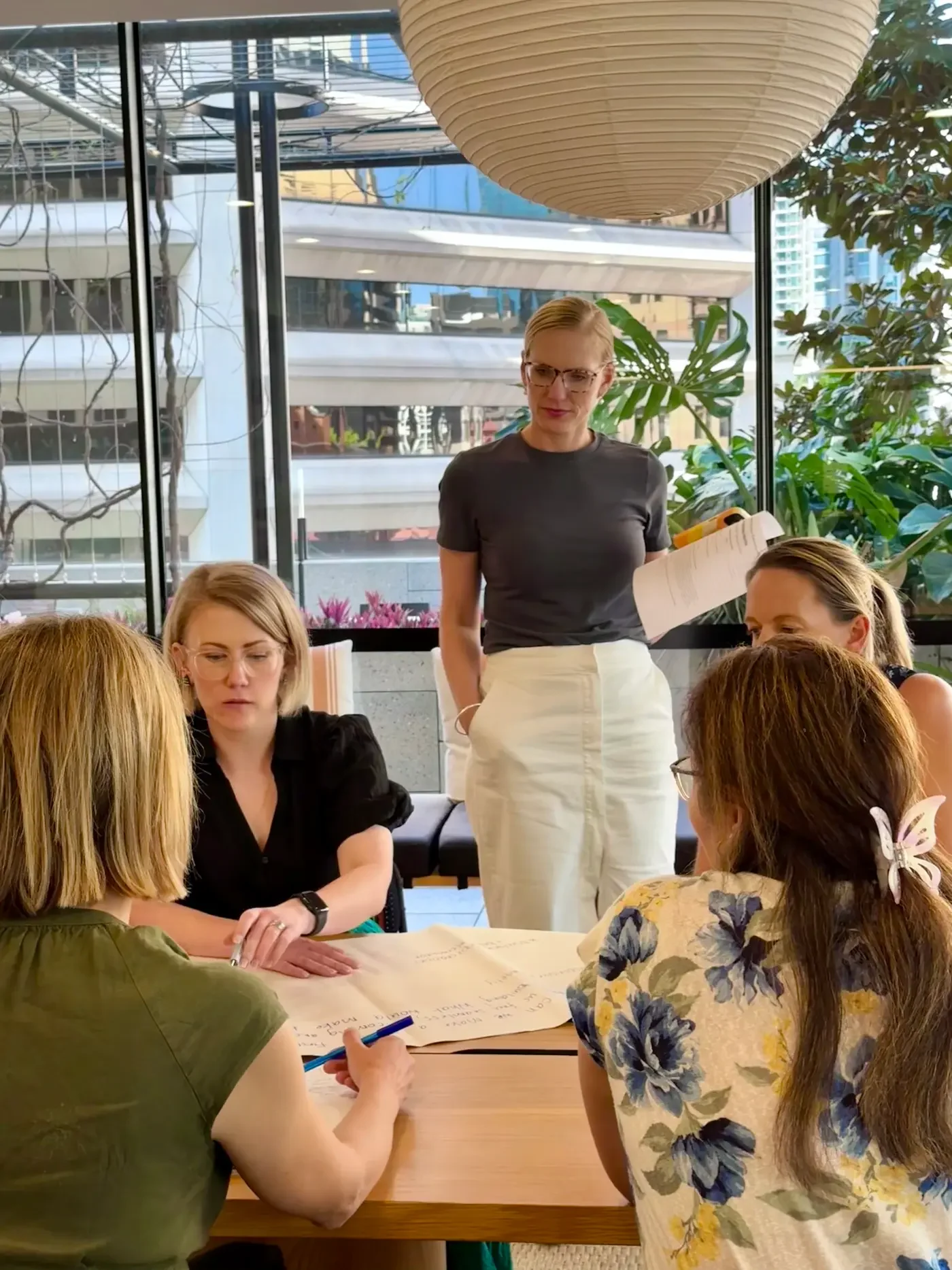 A group of women sitting around a table in a sunny room, engaging in a discussion or collaborative activity, with a woman standing and holding papers.