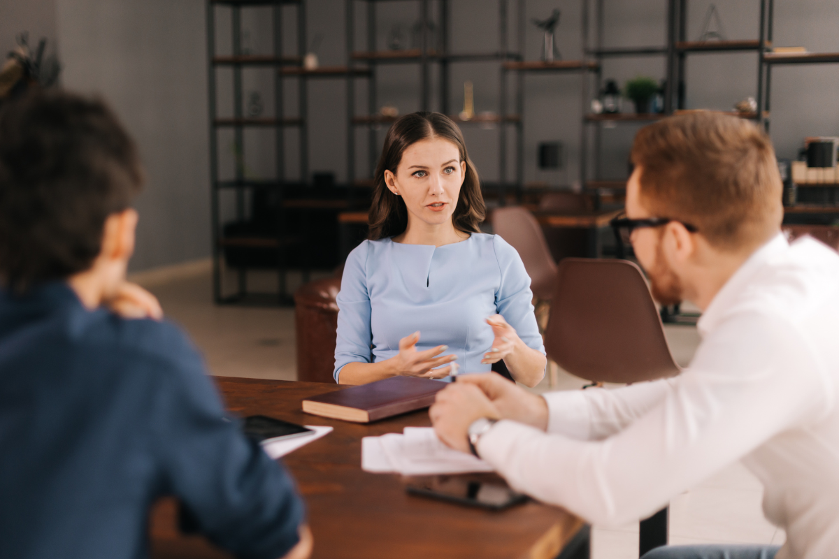 Three people having a discussion in a modern office setting, with a woman in a light blue shirt speaking and two men listening.