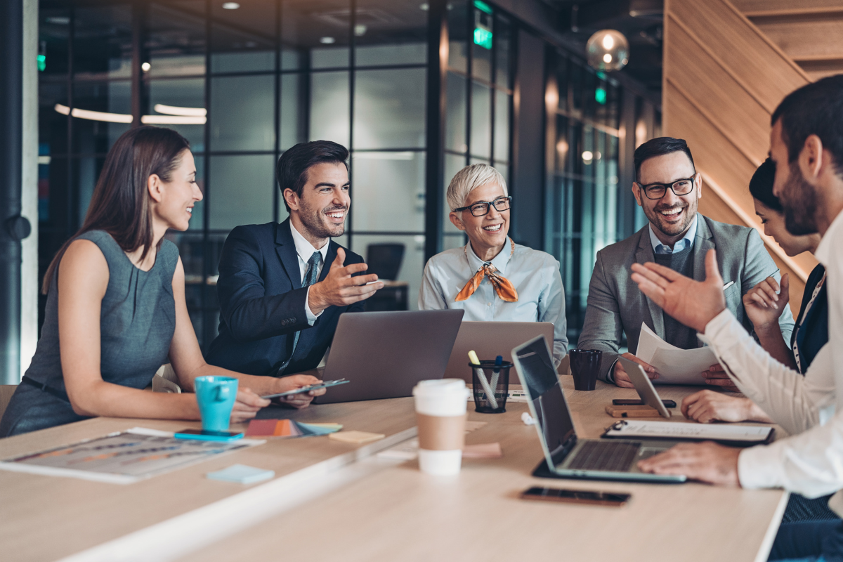 Group of six diverse professionals in business attire sitting around a conference table, smiling and engaging in conversation during a meeting in a modern office.
